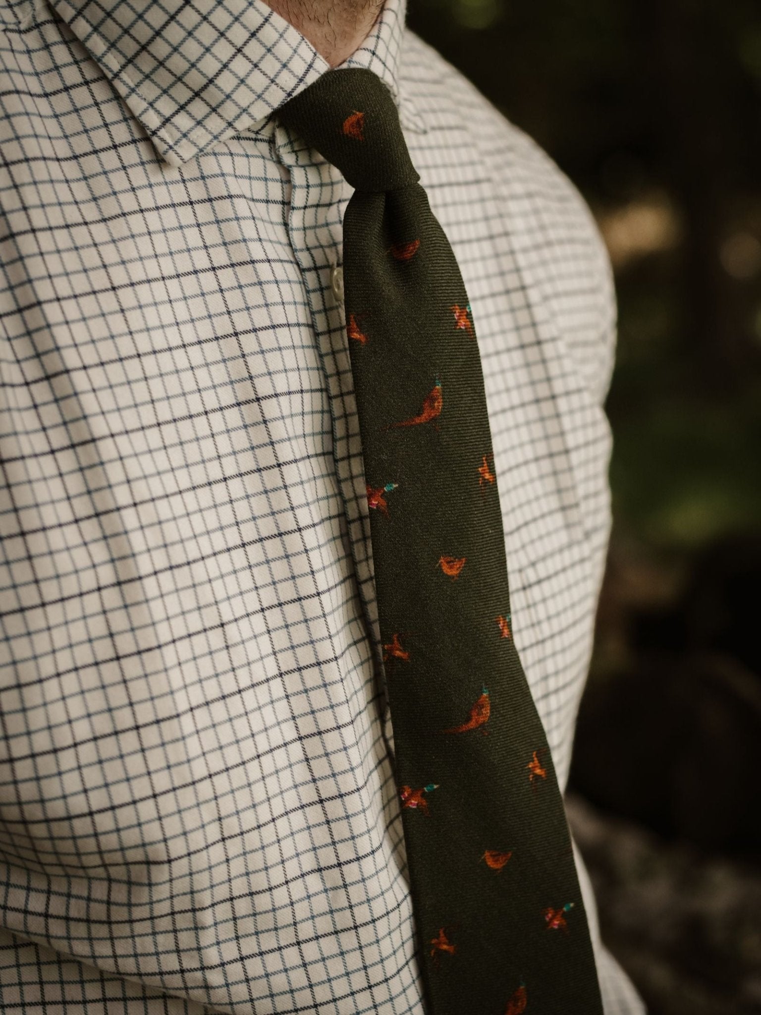 A person wears a white checkered button-up and the Campbell's of Beauly Mixed Game Silk Tie in dark green, featuring subtle countryside motifs. The image is cropped to show only the shirt, tie, and part of the neck.
