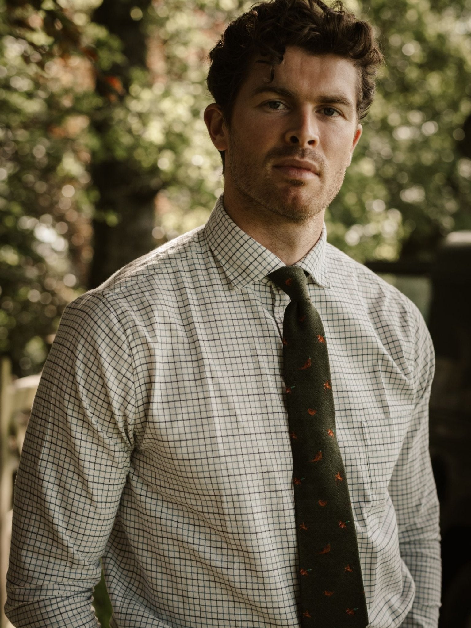 A man with short curly hair, in a checkered shirt and a Campbell's of Beauly Mixed Game Silk Tie, stands outdoors before blurred trees and sunlight.