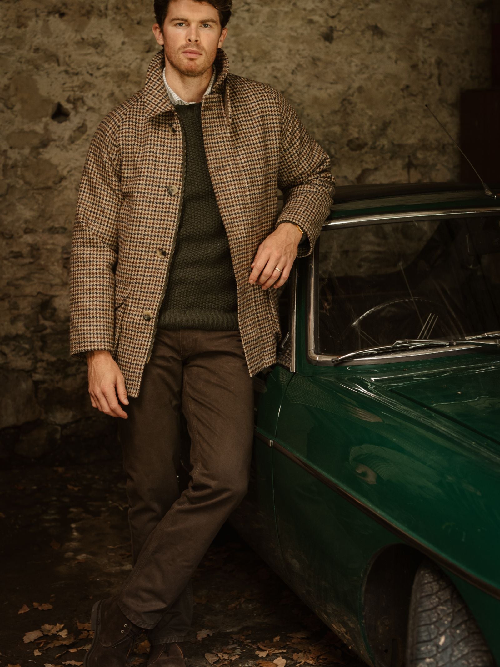 A man in Campbell's of Beauly Neutral Cotton Jeans leans against a vintage green car, with a brown checked jacket and green sweater, in front of a rustic stone wall.