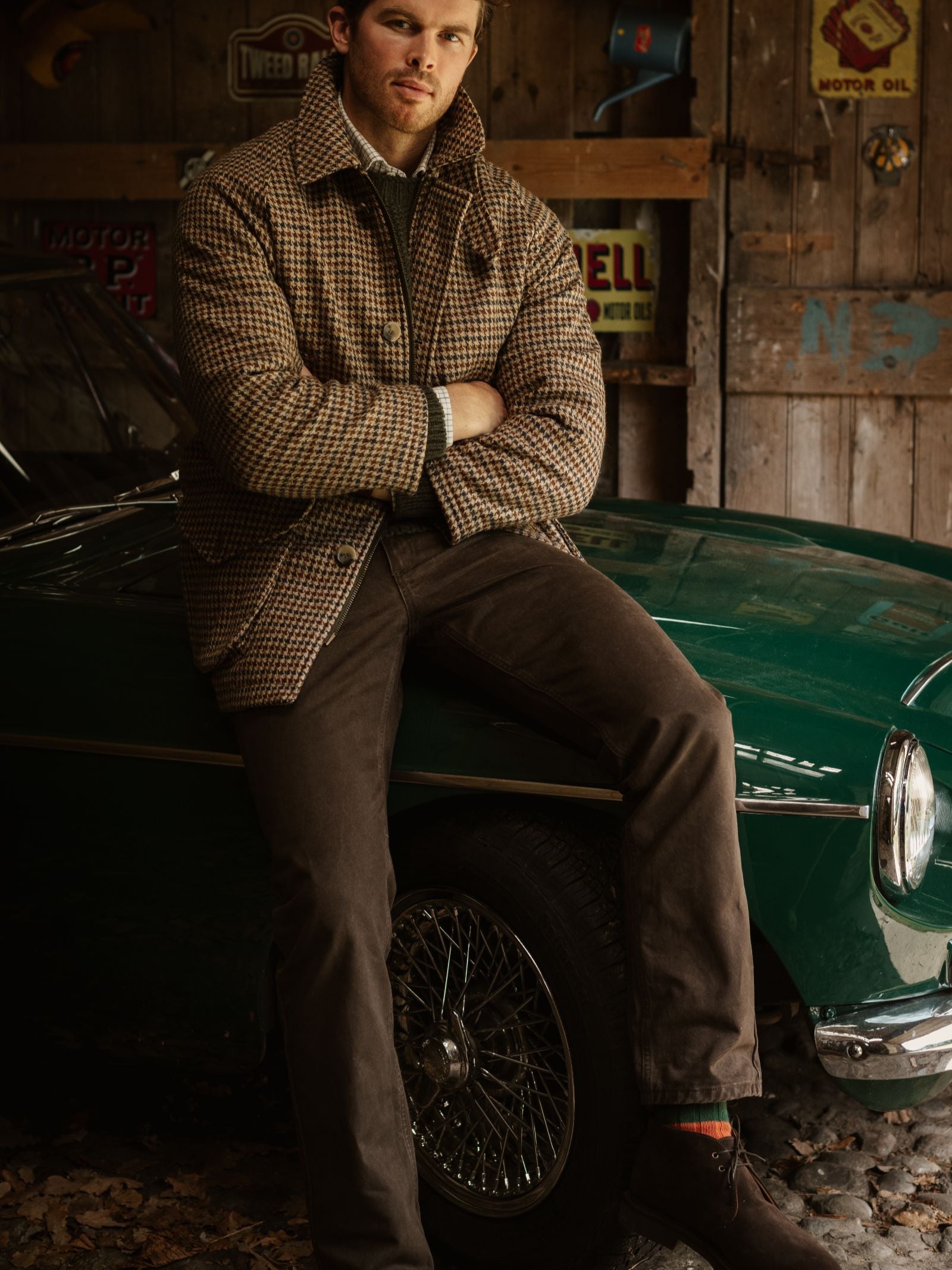 A man in a brown houndstooth jacket and Neutral Cotton Jeans by Campbell's of Beauly sits on the fender of a classic green car in a rustic garage, vintage signs lining the wooden wall behind him.