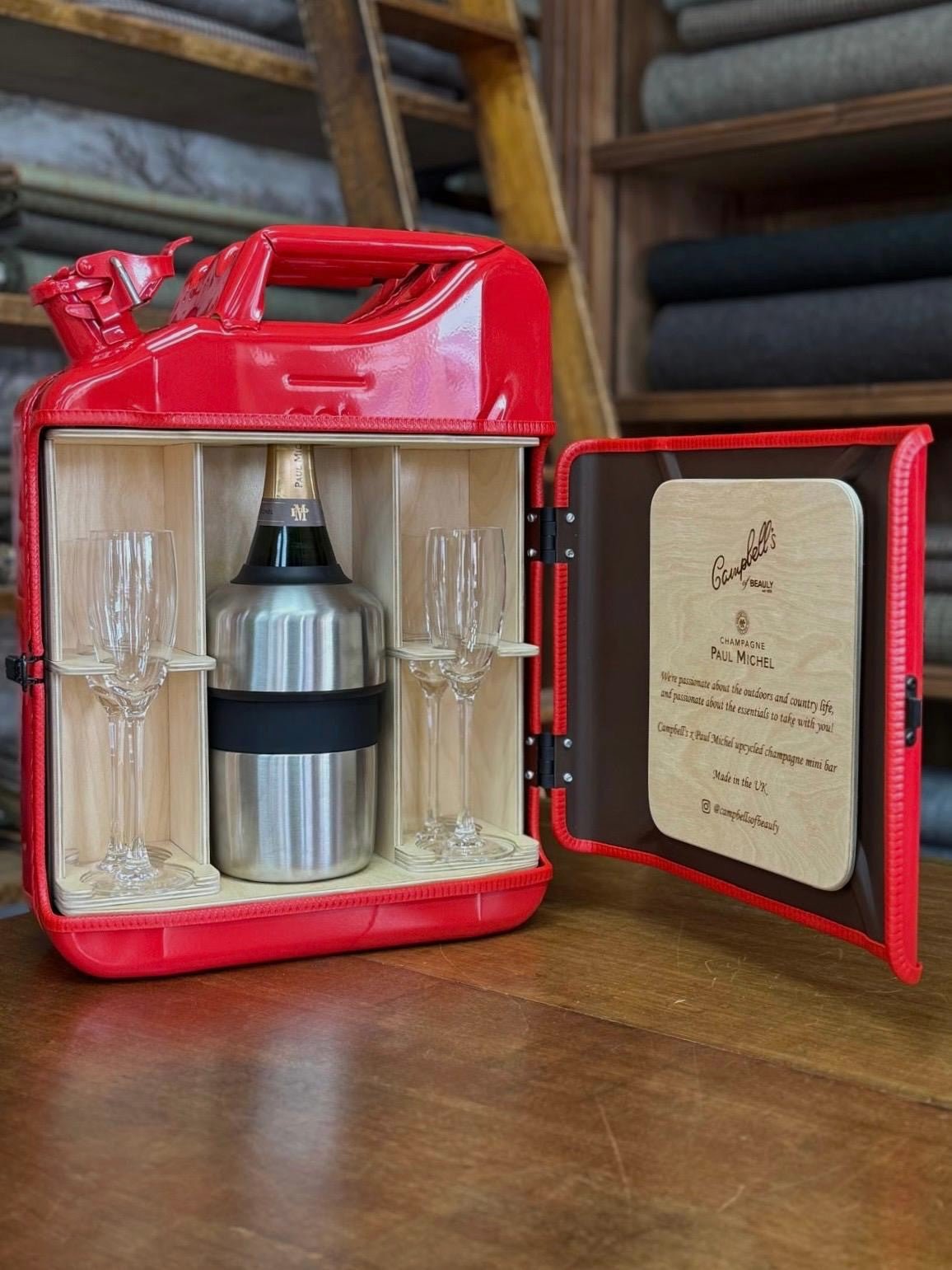 The Paul Michel Champagne Upcycled Mini Bar by Campbell's of Beauly transforms a red jerry can into a UK-crafted portable bar, holding champagne, two flutes, and a personalized plaque, displayed on wood.