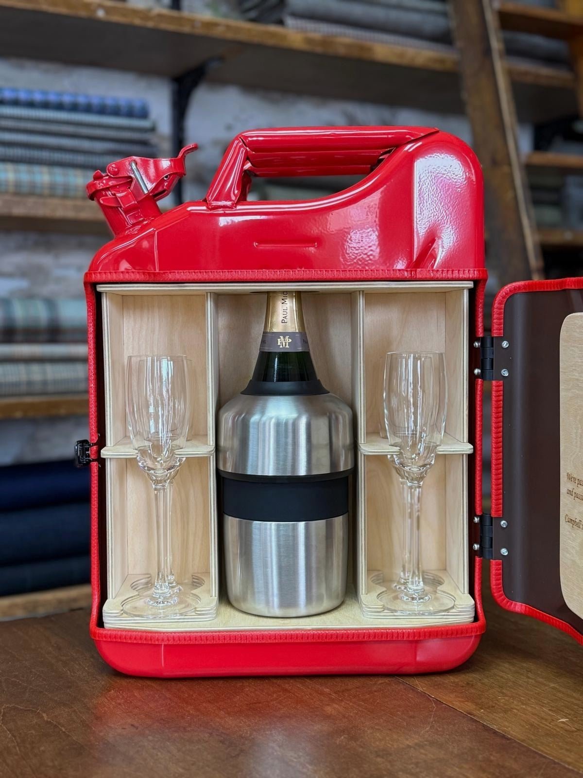 The Paul Michel Champagne Upcycled Mini Bar by Campbell's of Beauly is a red metal jerry can with a wooden interior, featuring space for a champagne bottle in a cooler and two flutes. It sits on a wooden table with fabric accents behind.
