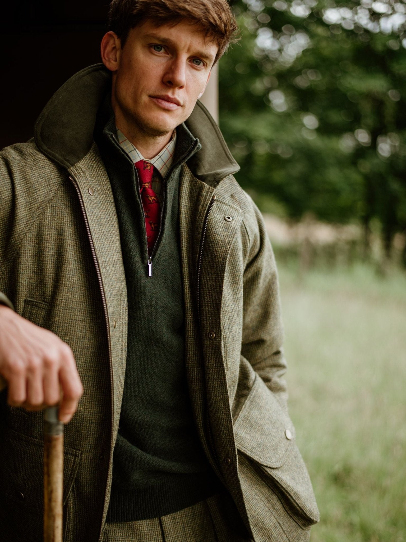 A man in country attire stands outdoors amid lush greenery, wearing a green jacket layered over the Campbell’s of Beauly Piquet Quarter Zip Jumper, paired with a plaid shirt and red tie, while holding a walking stick.