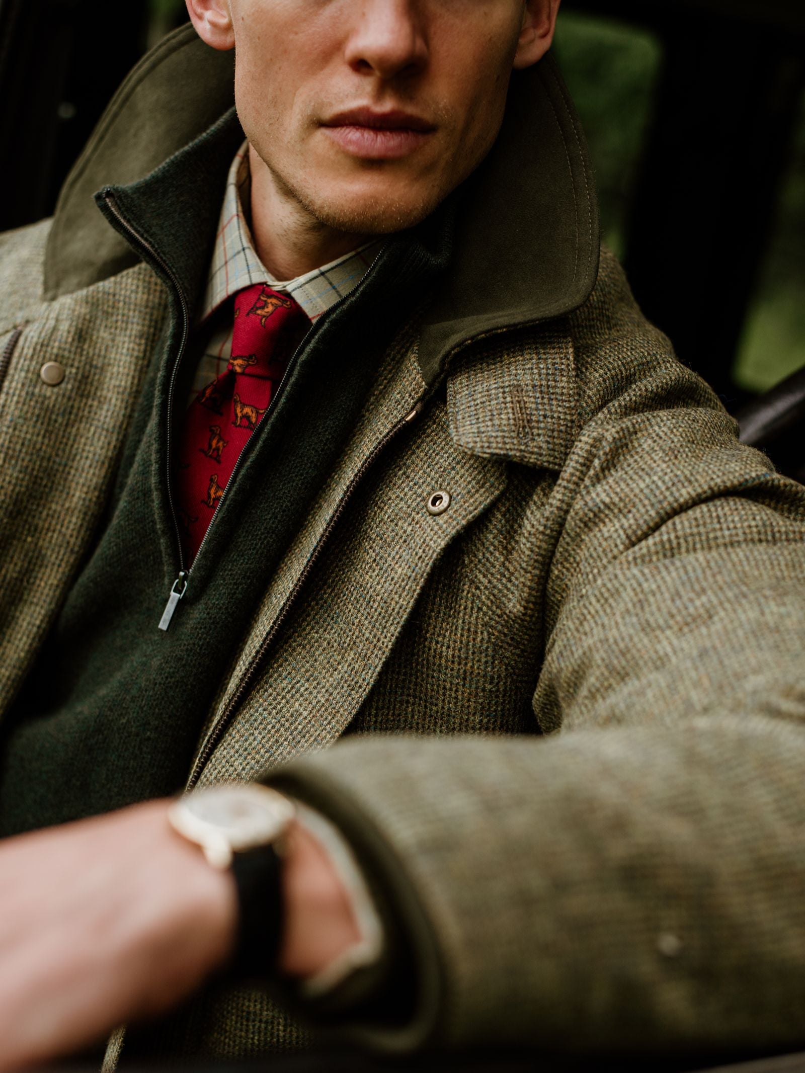 A man, seated in a car, wears the Campbell’s of Beauly Piquet Quarter Zip Jumper in green, layered over a plaid shirt and red patterned tie, with a tweed jacket on top and a watch on his wrist. His face is partially visible with a neutral expression.