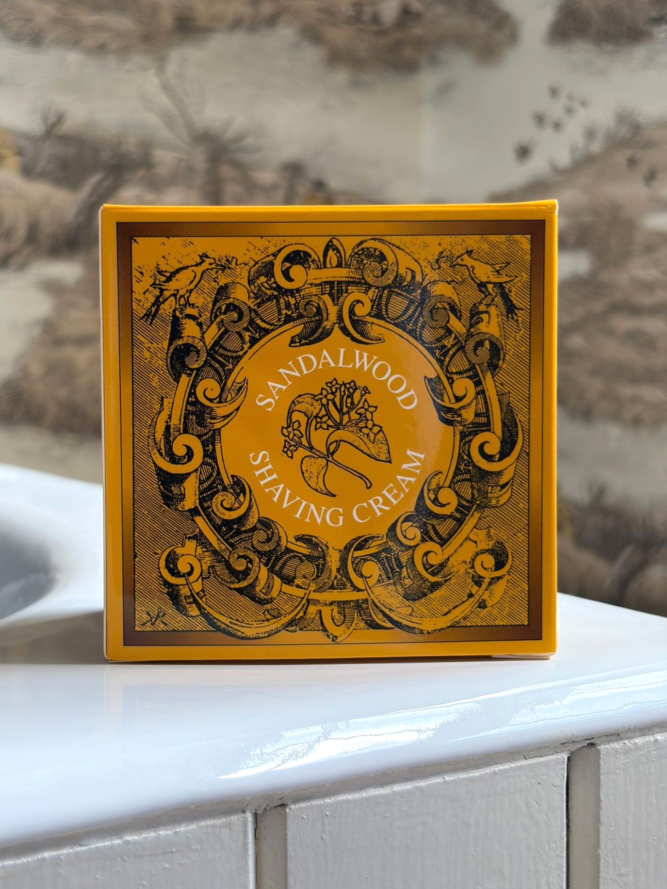 A yellow and brown box of Geo. F. Trumper Sandalwood Shaving Cream sits on white tiles by a sink, nature-themed wallpaper behind it—this gentle, glycerine-based cream is ideal for sensitive skin.