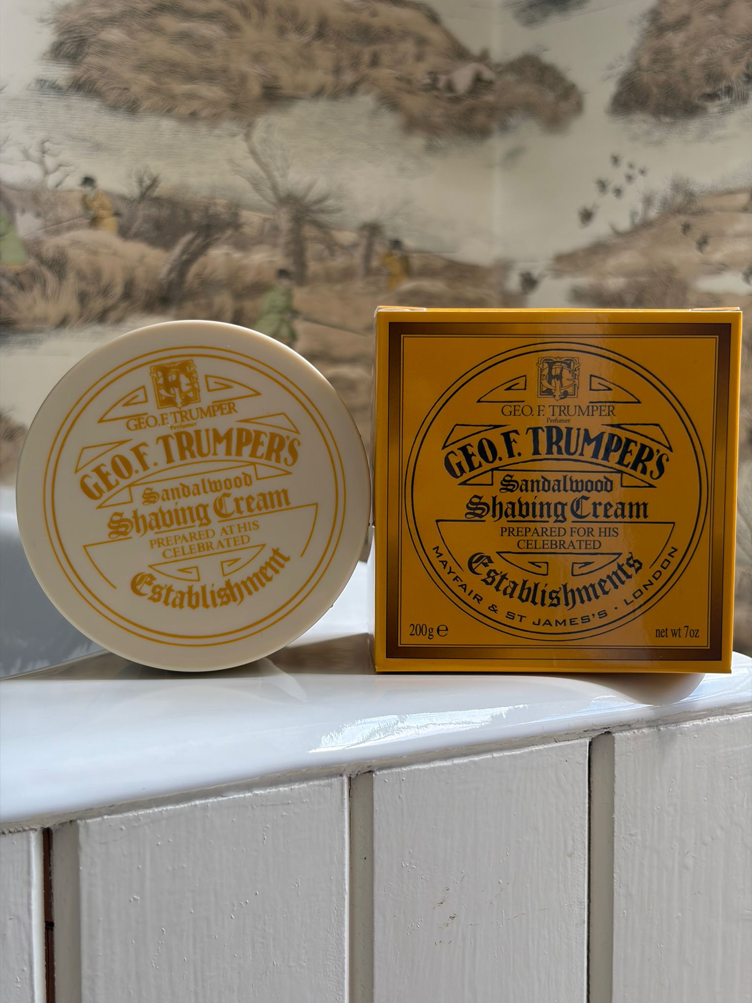 A white tub and yellow box of Geo. F. Trumper Sandalwood Shaving Cream rest on white tiles, set against animal-themed wallpaper.