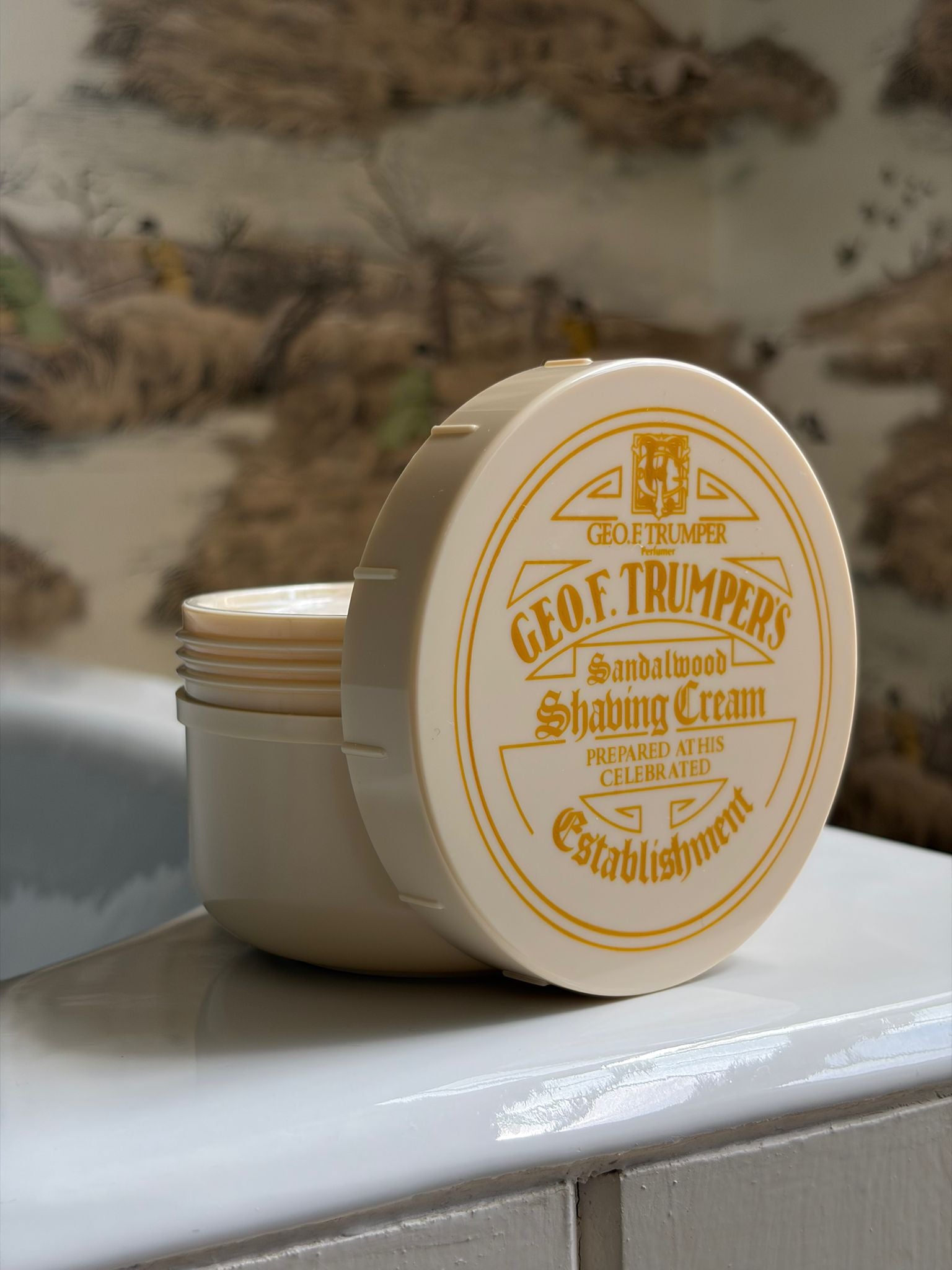 A round container of Geo. F. Trumper Sandalwood Shaving Cream with its lid open sits on a white bathtub’s edge, set against scenic vintage-style wallpaper.