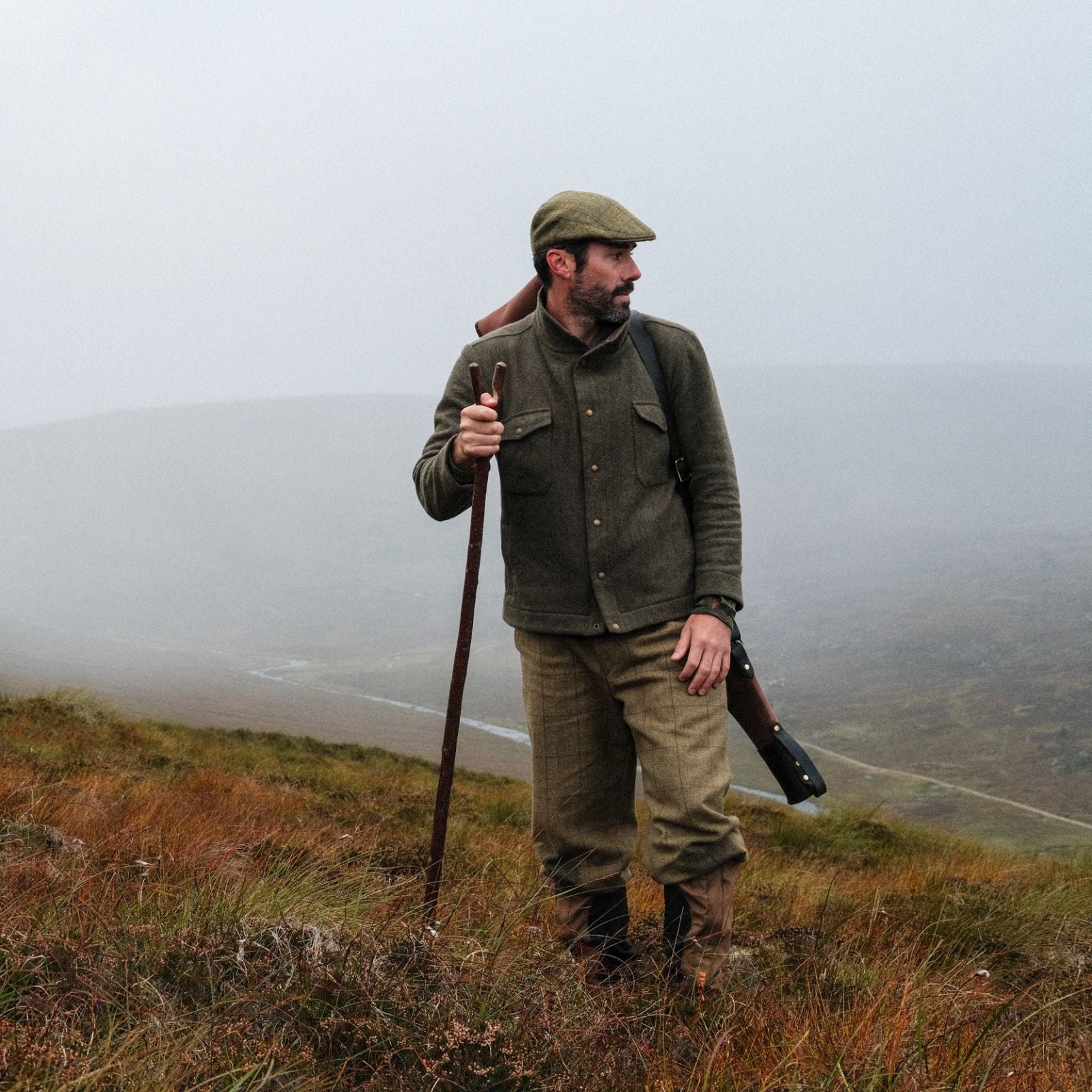A man in outdoor clothing stands on a misty hillside, holding a walking stick with a rifle slung over his shoulder. The landscape around him is grassy and slopes downward, with fog obscuring the background.