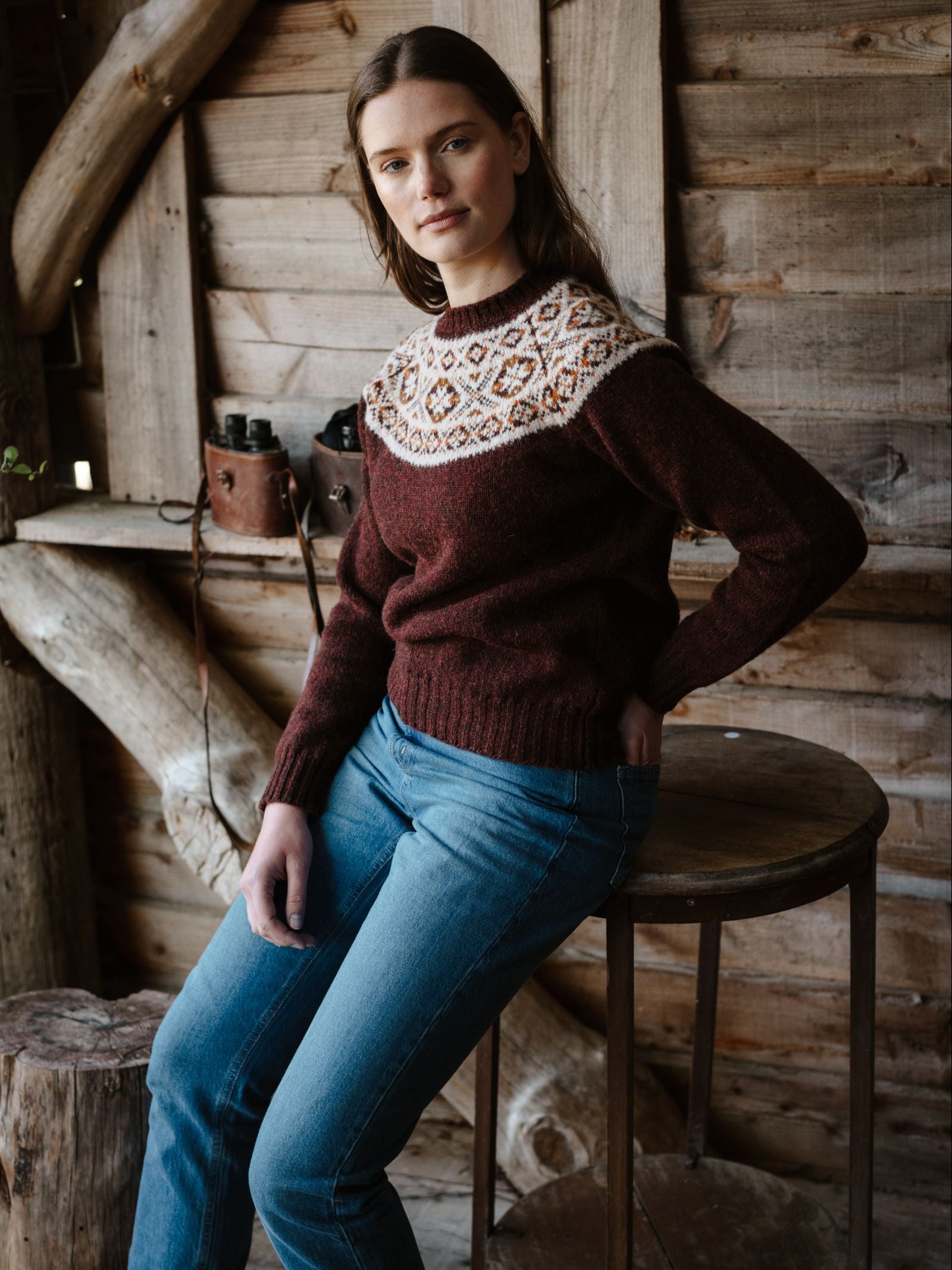 A woman with long brown hair wears the Campbell's of Beauly Shetland Fairisle Crew sweater in maroon and white, paired with blue jeans, while sitting on a round wooden stool in a rustic cabin. Binoculars rest on the shelf behind her.