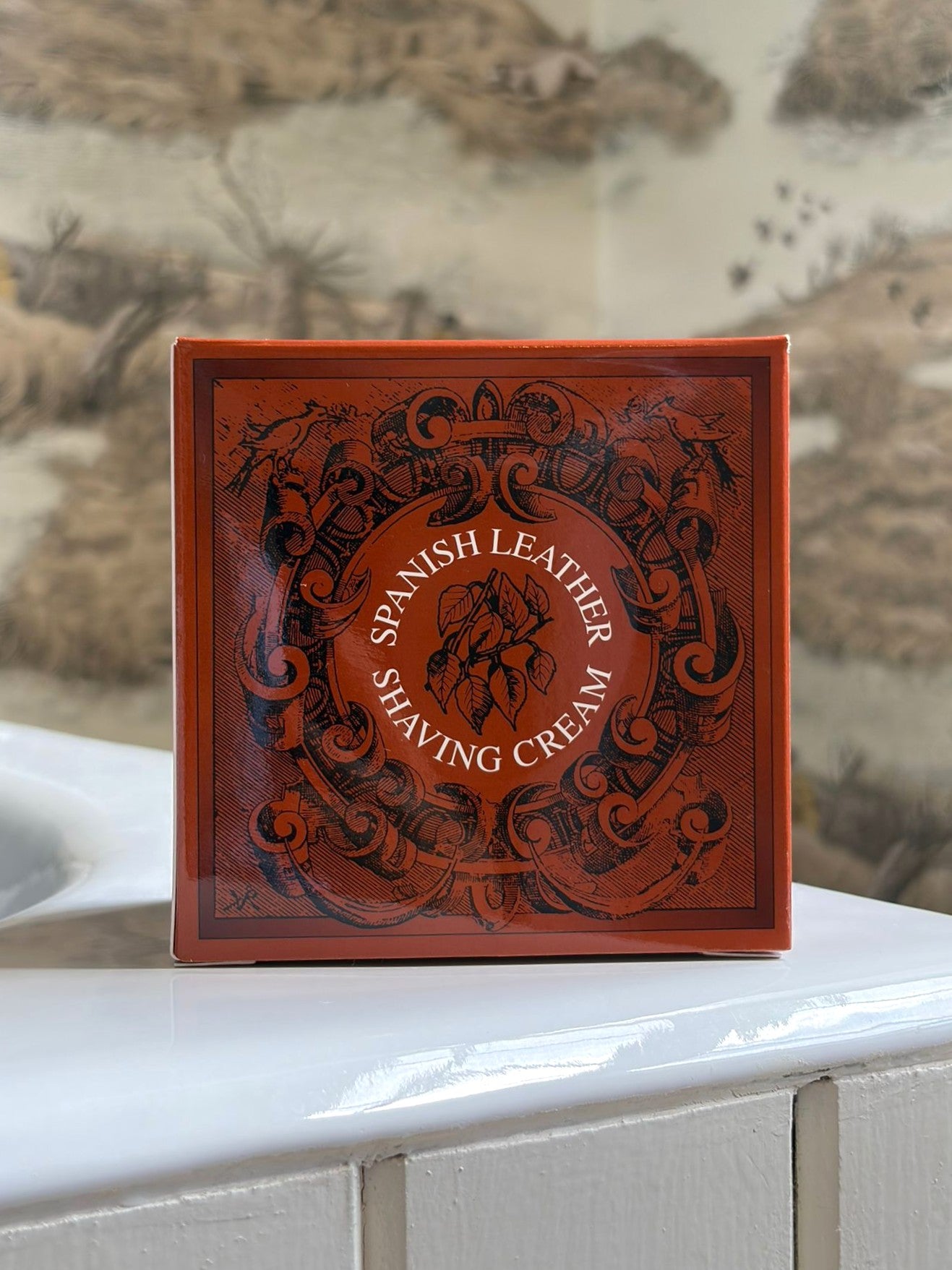A red box labeled Geo. F. Trumper Spanish Leather Shaving Cream 200g, featuring an ornate black design, sits on a white bathroom counter with blurred nature-themed wallpaper behind. This glycerine-based cream is ideal for sensitive skin.