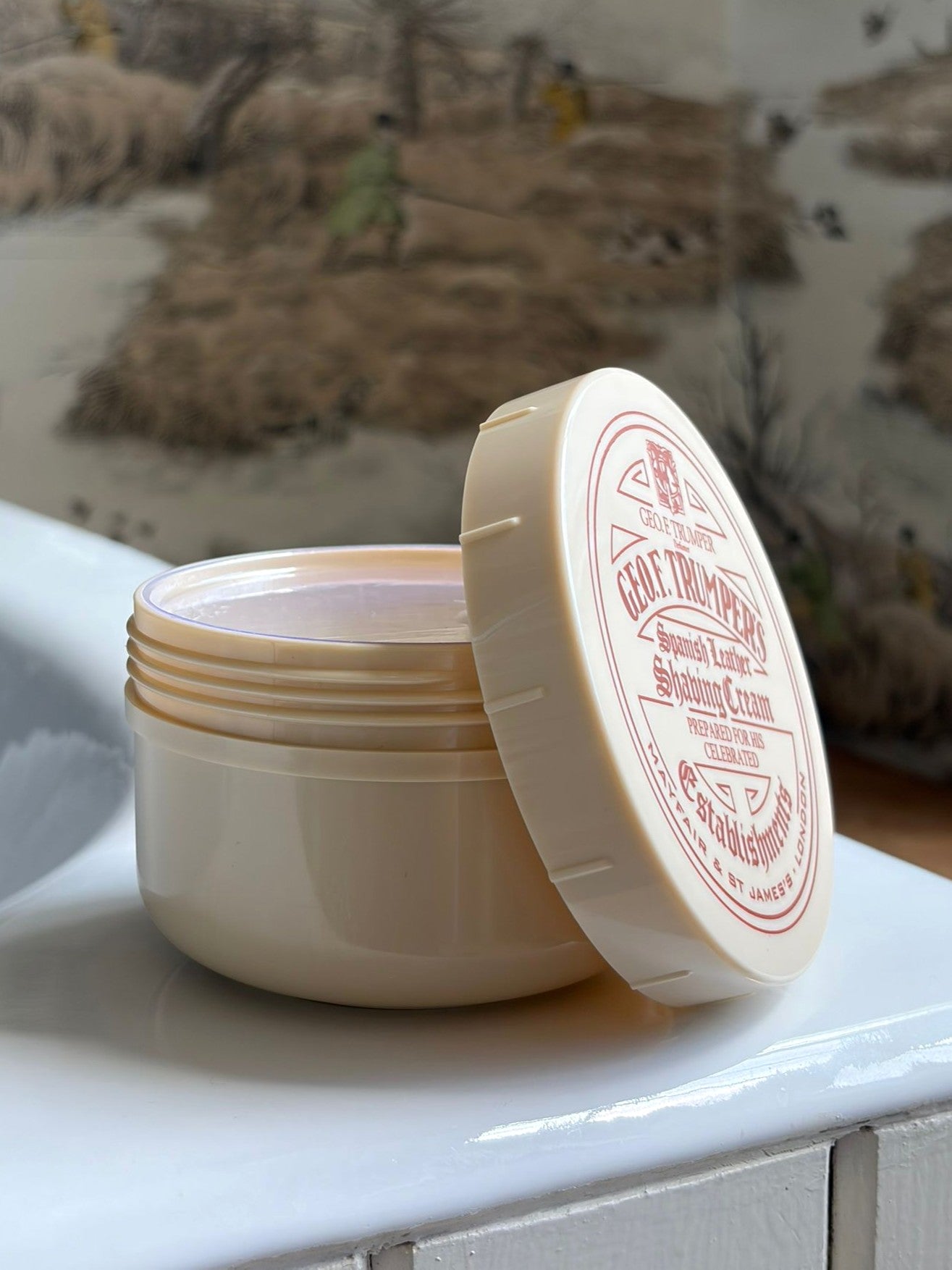 An open container of Geo. F. Trumper Spanish Leather Shaving Cream 200g sits on a white surface with its lid nearby, while a blurred, patterned wall in the background evokes a luxurious shaving experience.