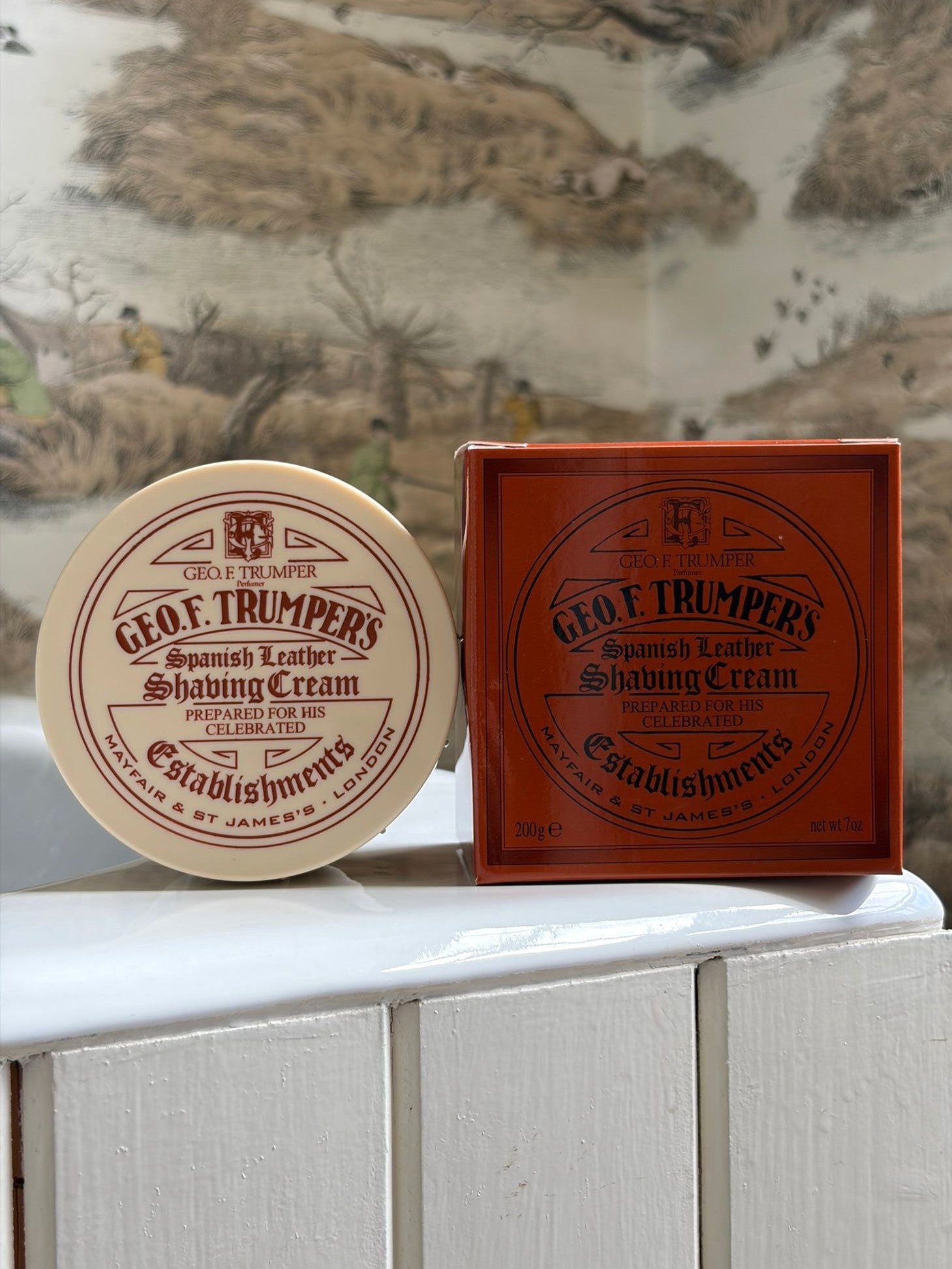 A round container and a square box of Geo. F. Trumper Spanish Leather Shaving Cream 200g sit on a white wooden surface with blurred, nature-inspired wallpaper in the background—ideal for a luxurious shave.