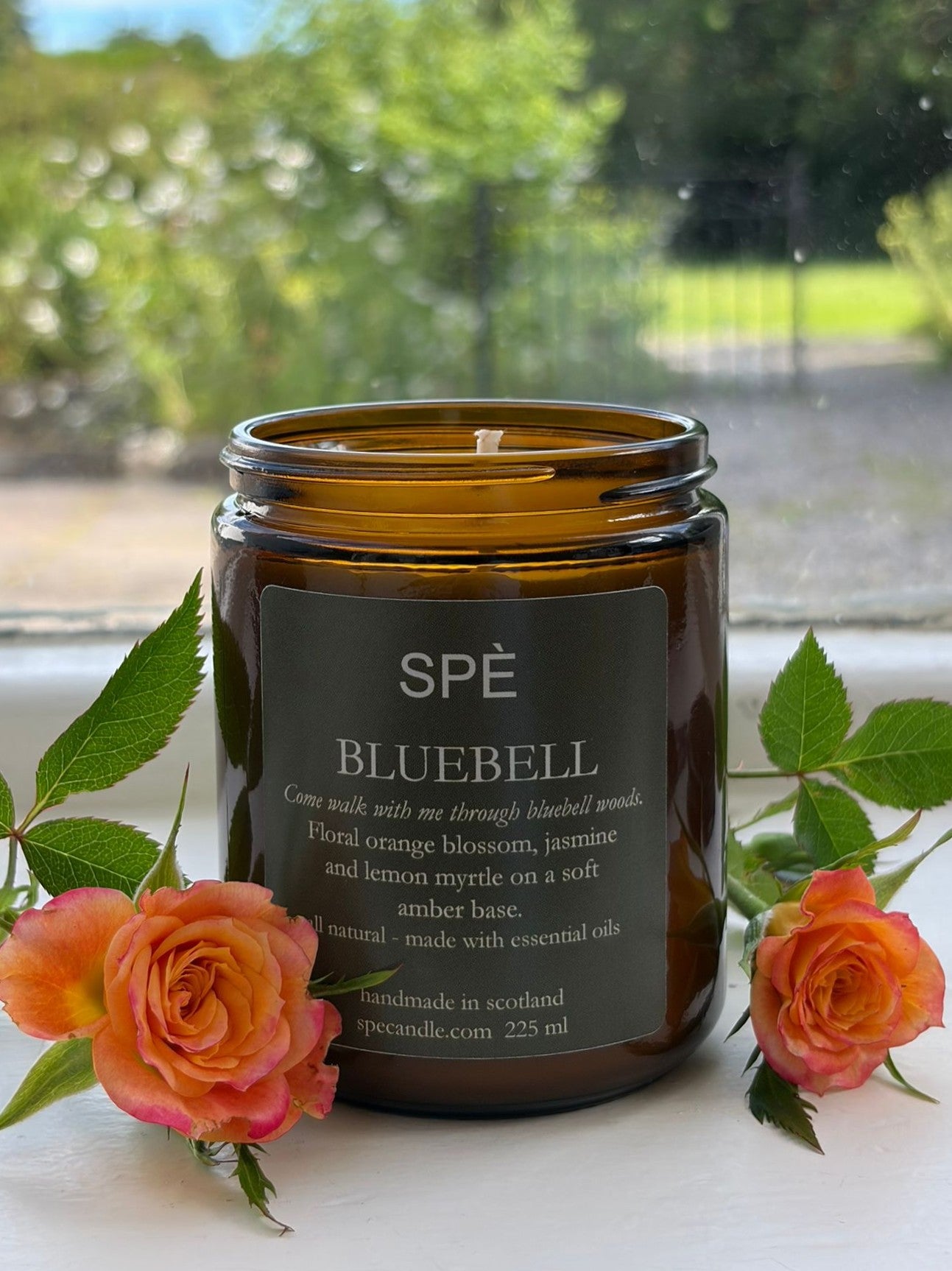 A Bluebell Candle by SPÈ Atelier, presented in an amber jar and labeled BLUEBELL, rests on a windowsill next to delicate pink roses. Beyond the window, a green sunlit garden and clear sky evoke the fresh woodland fragrance of spring with every gentle breeze.