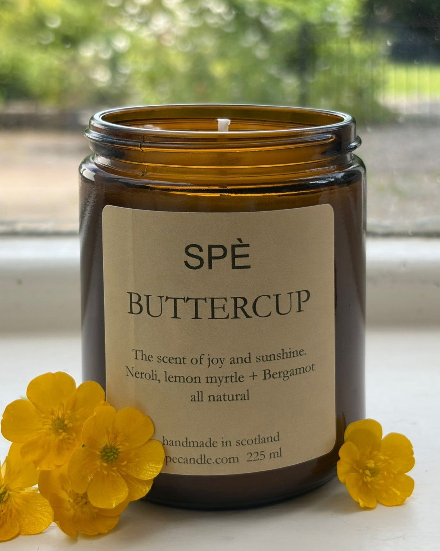 A brown glass jar candle labeled “Buttercup Candle” by SPÈ Atelier rests on a windowsill, surrounded by yellow buttercup flowers. Its label highlights notes of neroli, lemon myrtle, and bergamot, creating a vibrant citrus floral fragrance accented by a delicate hint of buttercup.