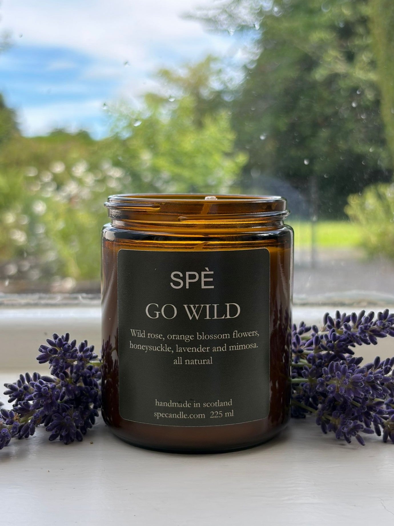 A brown glass candle labeled SPÈ Atelier Go Wild Candle rests on a windowsill, surrounded by sprigs of lavender and lush green botanicals. Through the window, a verdant garden and blue sky can be seen. The setting radiates soft floral notes and natural aromatics from the candle, creating a serene and inviting atmosphere.