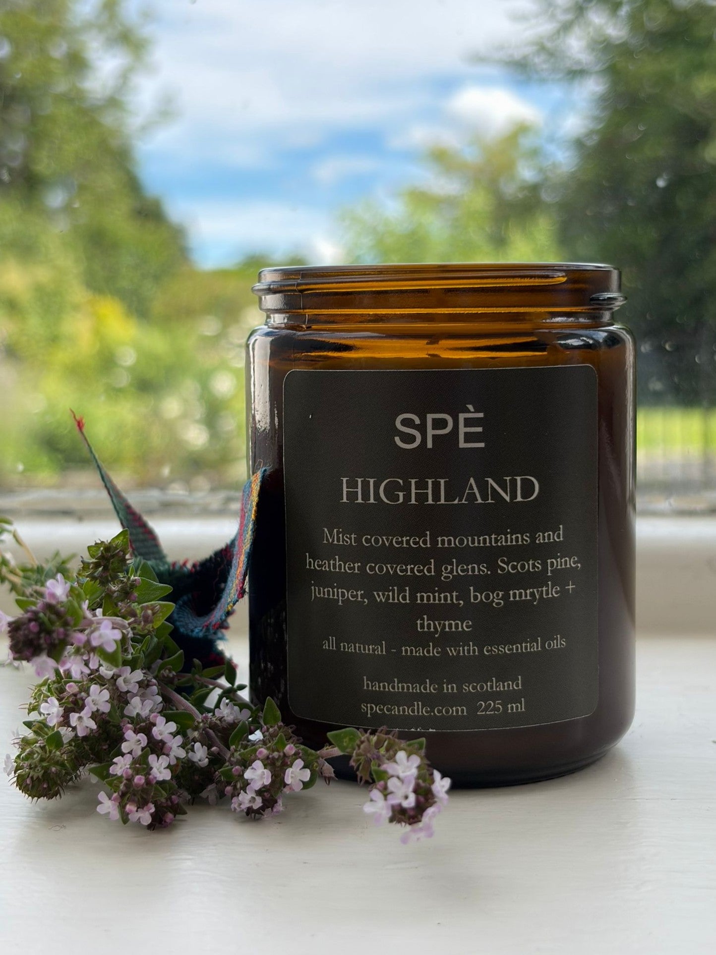 A brown glass Highland Candle by SPÈ Atelier rests on a windowsill beside small purple flowers. Through the window, trees and grass are visible, enhancing the botanical fragrance inspired by the Scottish Highlands with gentle hints of juniper.