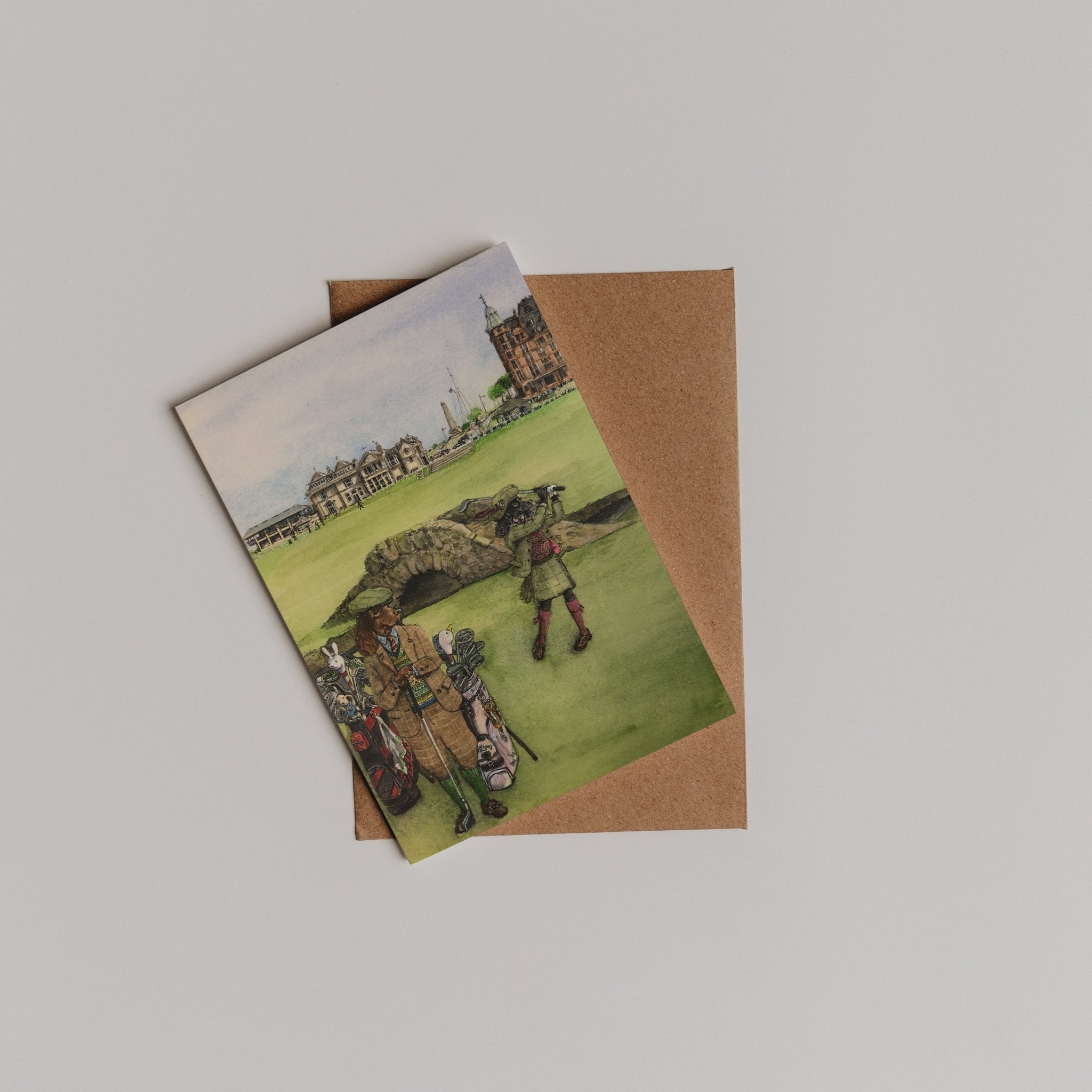 A folded brown envelope with a thank you card on top, the St Andrews Swilken Bridge Greeting Card by Campbell’s of Beauly, featuring people in vintage clothing on a lawn before historic buildings and the iconic stone bridge.
