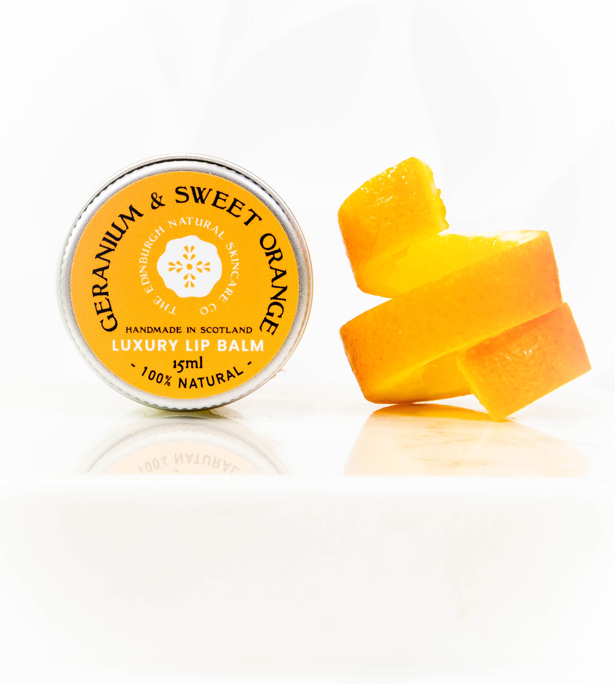 A tin of The Edinburgh Natural Skincare Company’s Geranium & Sweet Orange Luxury Lip Balm rests beside a twisted orange peel on a white background. The yellow-labeled balm is handmade in Scotland.
