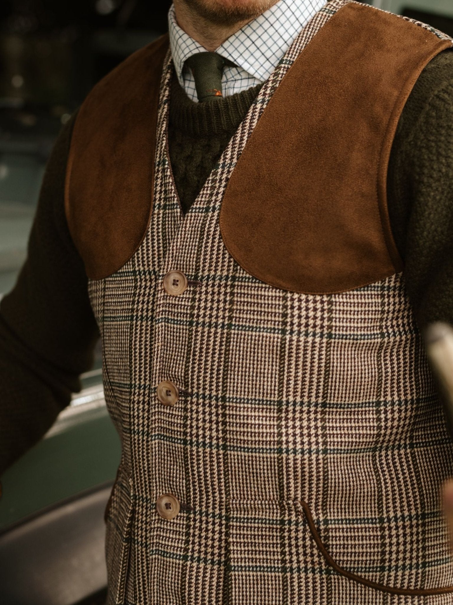 A person wearing the Campbell's of Beauly Tweed Shooting Waistcoat with suede shoulder panels over a dark green sweater and checkered shirt, paired with a dark green tie—ideal for countryside pursuits.