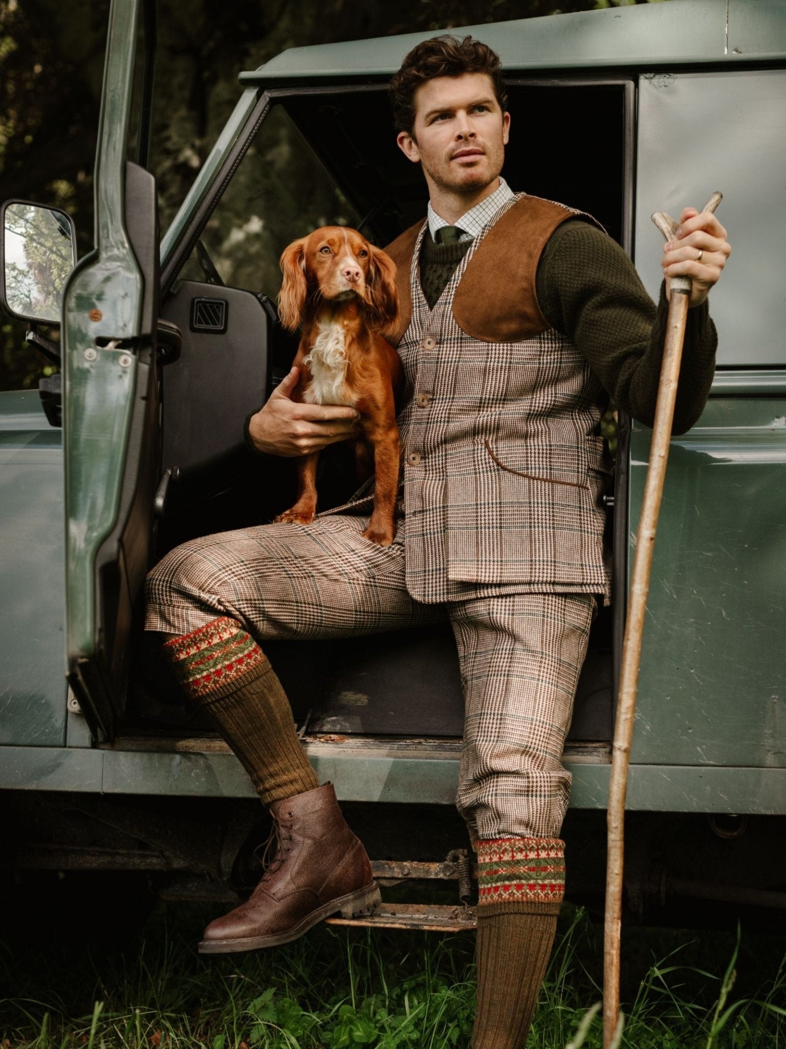 A man in a Campbell's of Beauly Tweed Shooting Waistcoat sits by an open vehicle door, holding a brown and white dog and walking stick, enjoying greenery—a perfect countryside pursuits moment.