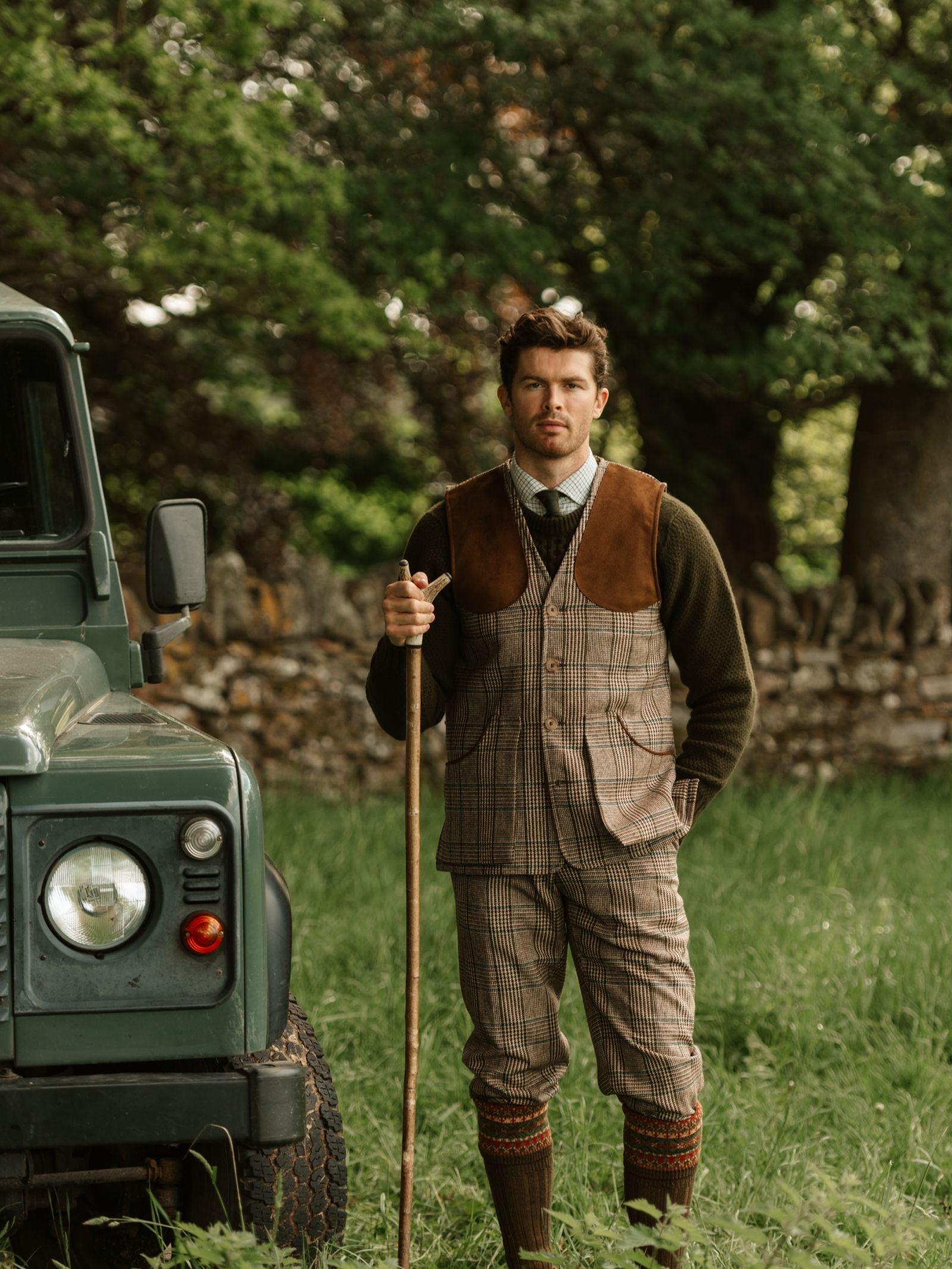 A man in traditional hunting attire and a Campbell's of Beauly Tweed Shooting Waistcoat stands by a green off-road vehicle on grass, holding a wooden walking stick, with trees and a stone wall behind—ideal for countryside pursuits.