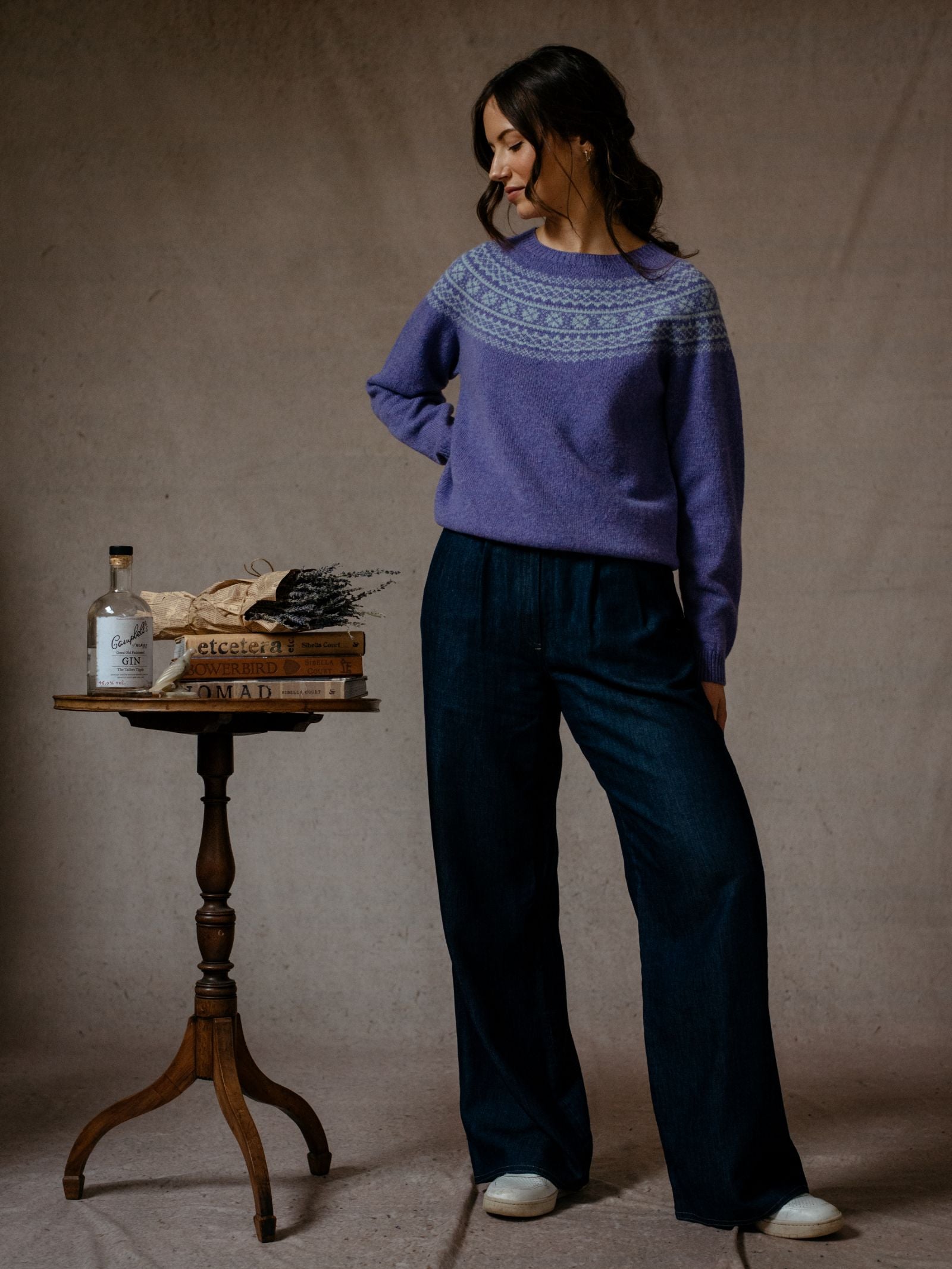 A woman in Campbell's of Beauly's Auld Stock Two-Colour Fairisle Yoke Jumper and wide-leg dark jeans stands by a small wooden table with books, a bottle, bread, and dried flowers. She smiles softly against a neutral background.