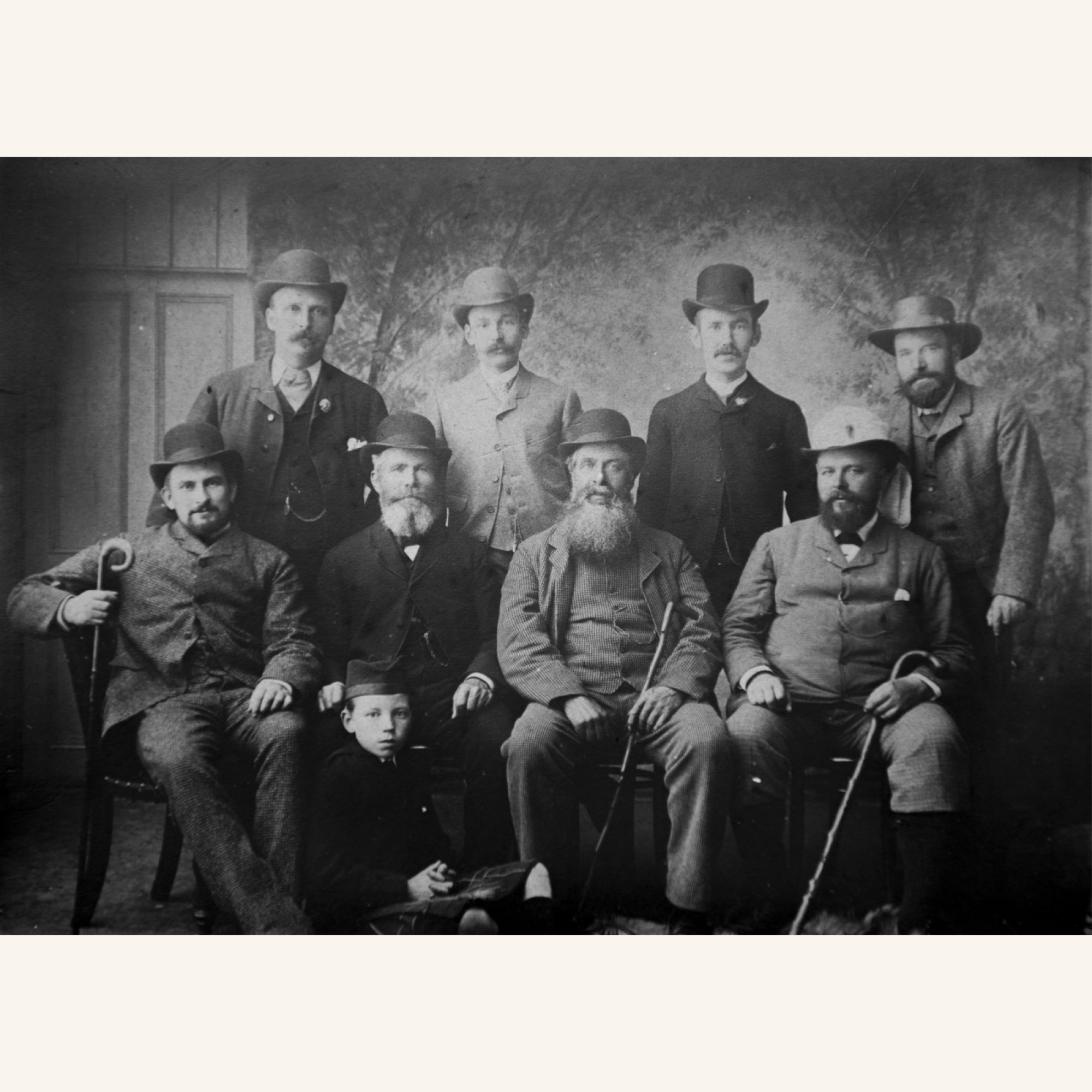 A black-and-white vintage photo of ten men, most with beards and mustaches, wearing suits and hats. Nine men are seated or standing, while one young boy sits cross-legged on the floor at the front.