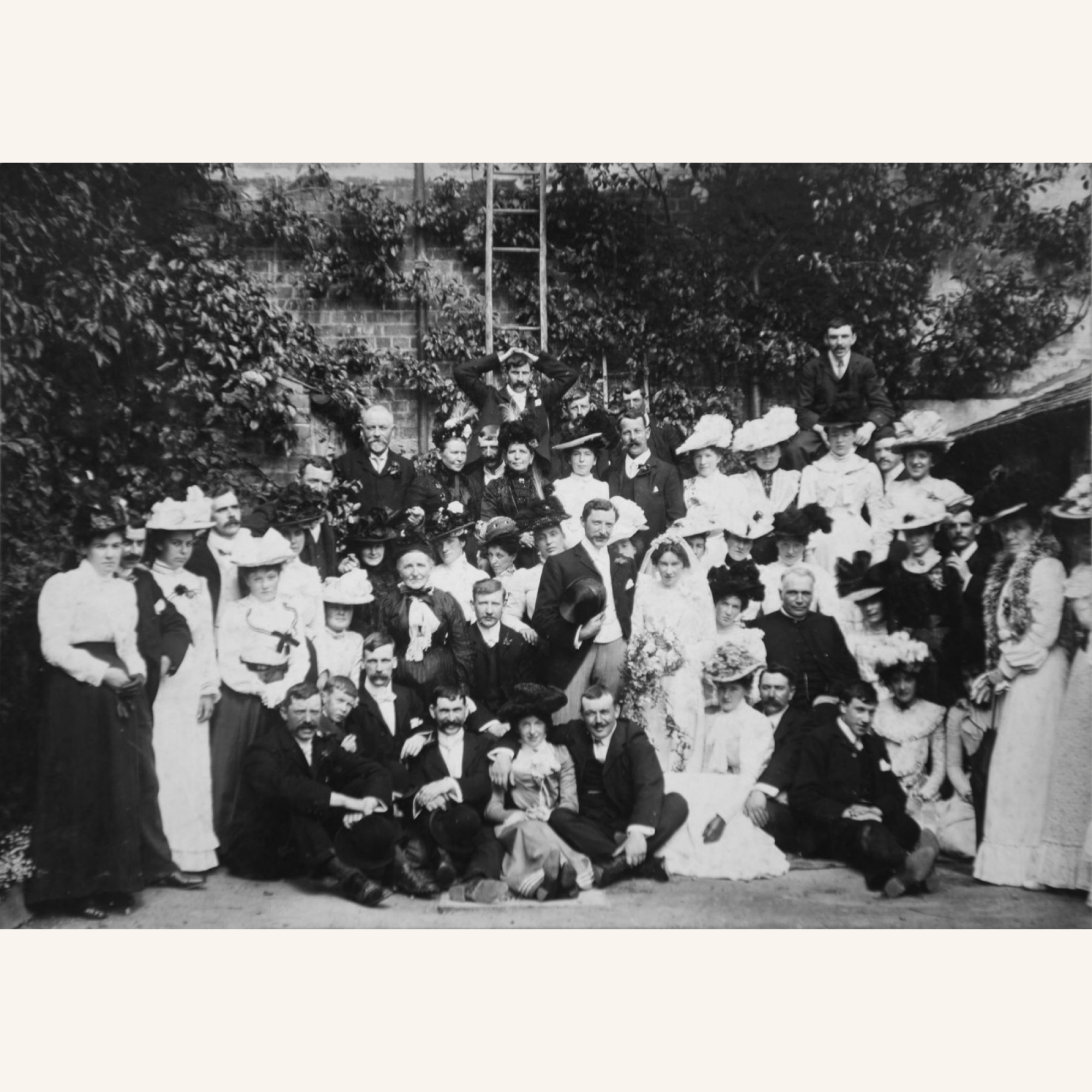 Black and white vintage photo of a large group of people, men and women in formal attire, posing outdoors in front of greenery. Some women wear hats and a bride is seated in the center, suggesting a wedding gathering.