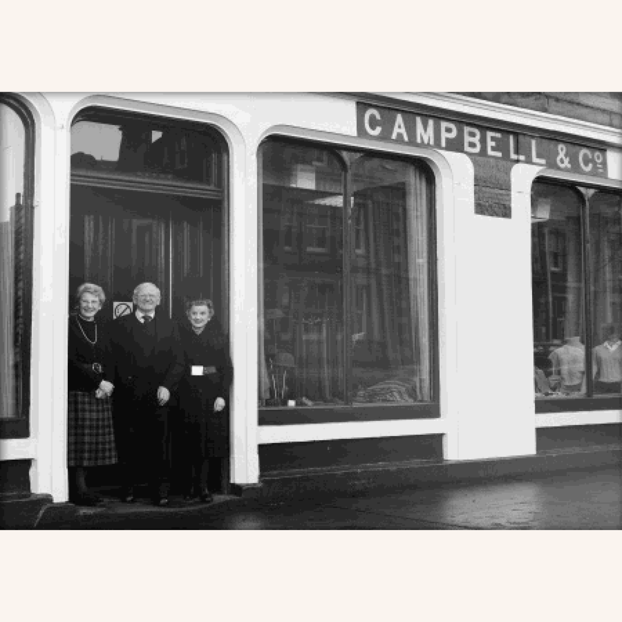 Three people, two women and one man, stand smiling in front of a shop with large windows and a sign above reading CAMPBELL & CO. The shop displays clothing inside. The photo is in black and white.