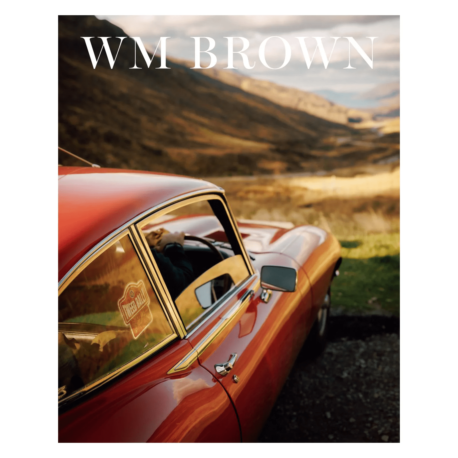 A classic red car is parked on a scenic countryside road with rolling hills, featured on the cover of William Brown Issue No 19. The WM Brown magazine title appears at the top in bold white letters.