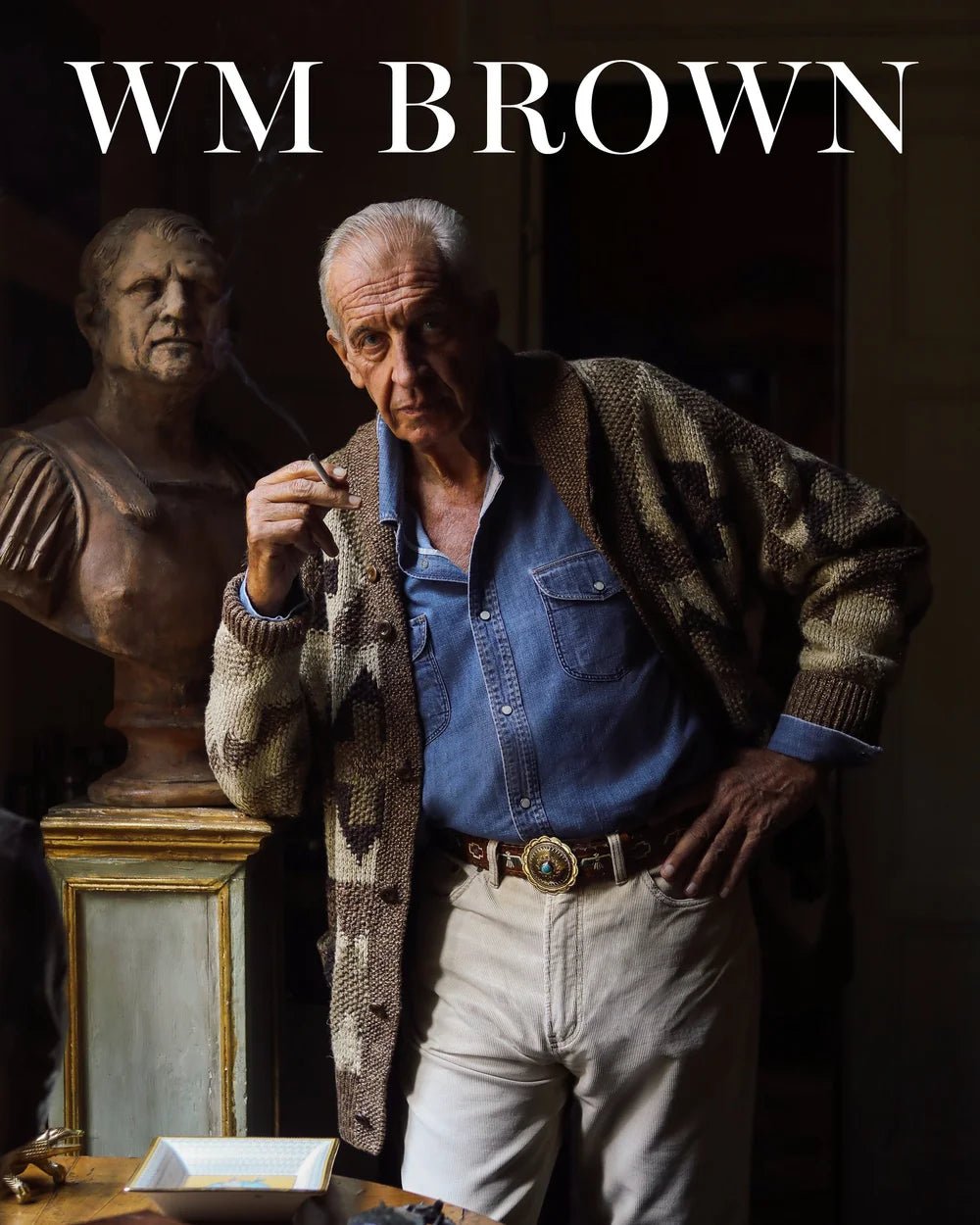 An older man in a denim shirt, patterned cardigan, and white pants leans on a table with a cigar beside a classical bust, expressing timeless style on the cover of WM Brown’s William Brown Issue No 21.
