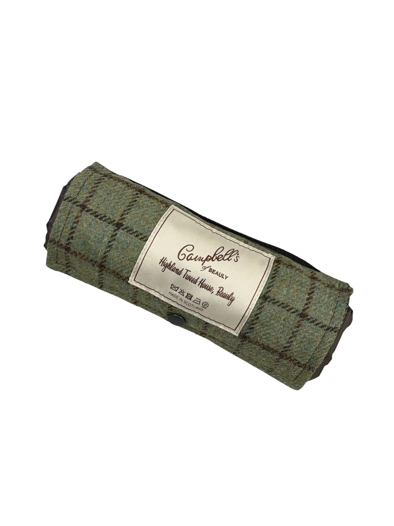 The Walker Camper Picnic Rug by Campbell’s of Beauly is a rolled-up green plaid fabric pouch featuring a front label with the brand name and “Highland Tweed House, Beauly.” Crafted from premium British wool, this classic picnic rug also boasts a practical waterproof backing.