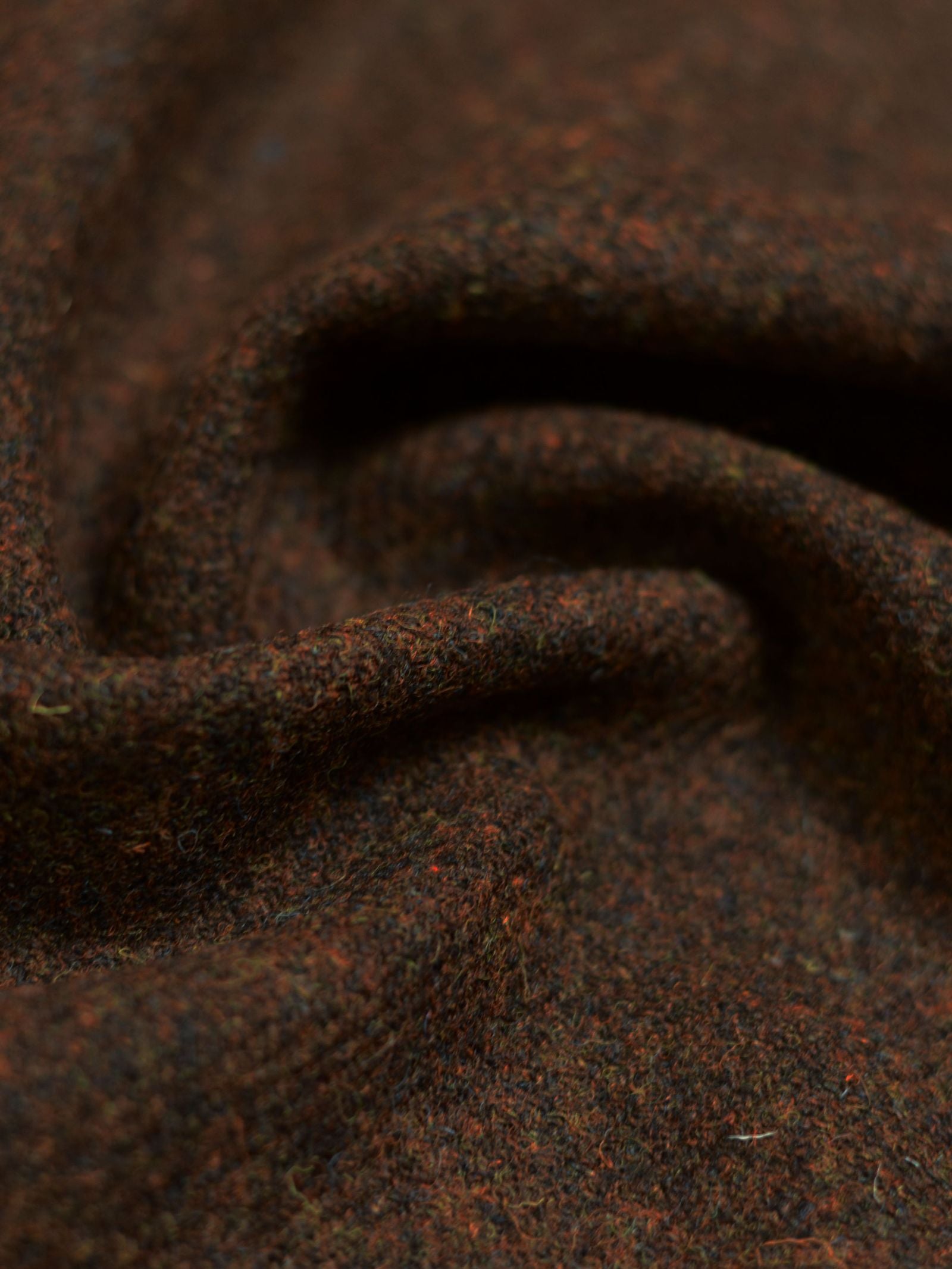 Close-up of Campbell's of Beauly Harris Tweed 93893 wool fabric in textured dark brown, featuring subtle red and green tones, displayed in gentle folds under soft lighting.