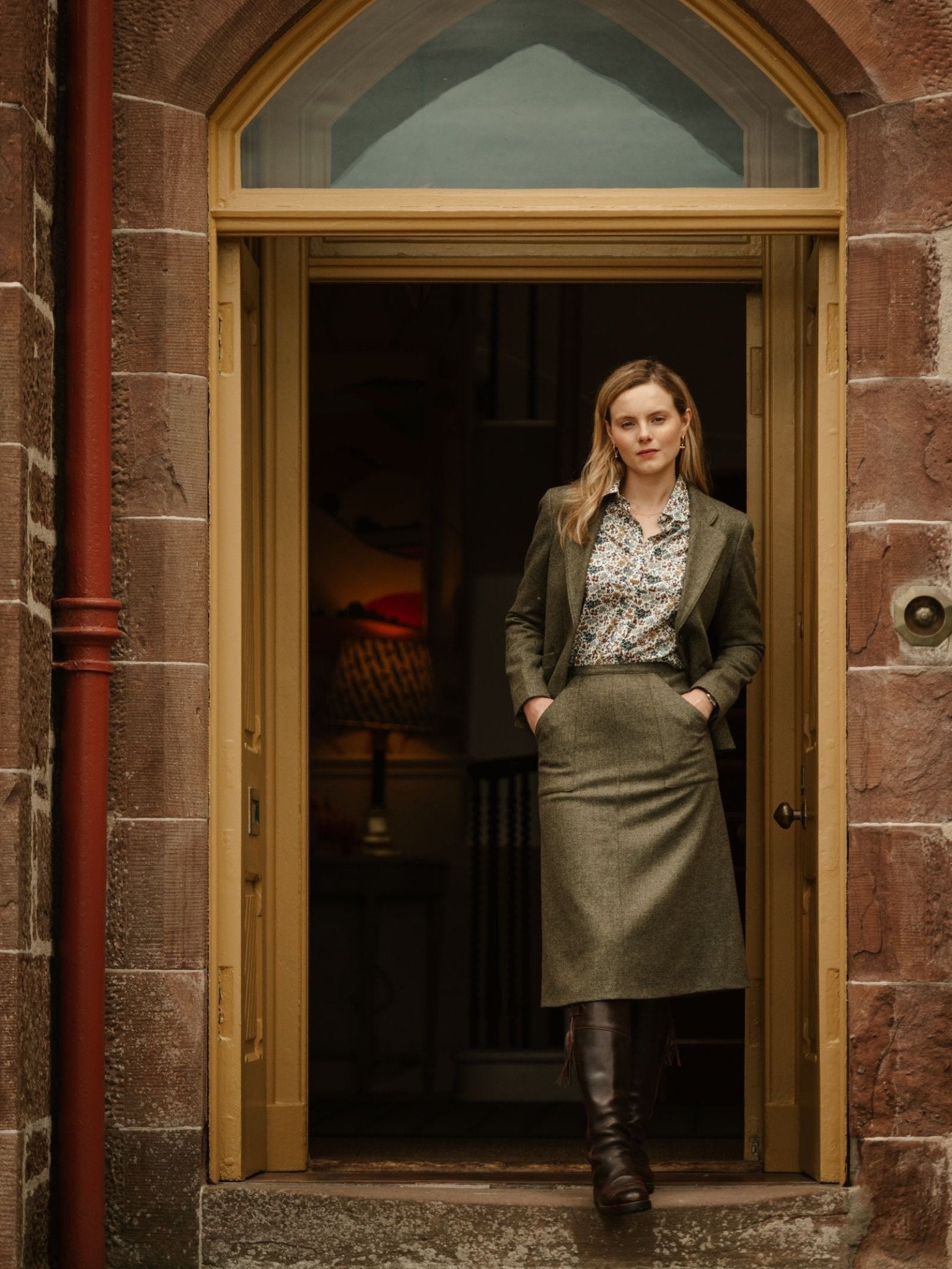 A woman stands in a stone doorway, hands in pockets, wearing a green blazer, floral blouse, Campbell's of Beauly A-Line Skirt, and brown knee-high boots; her long blonde hair frames her face as she looks at the camera.