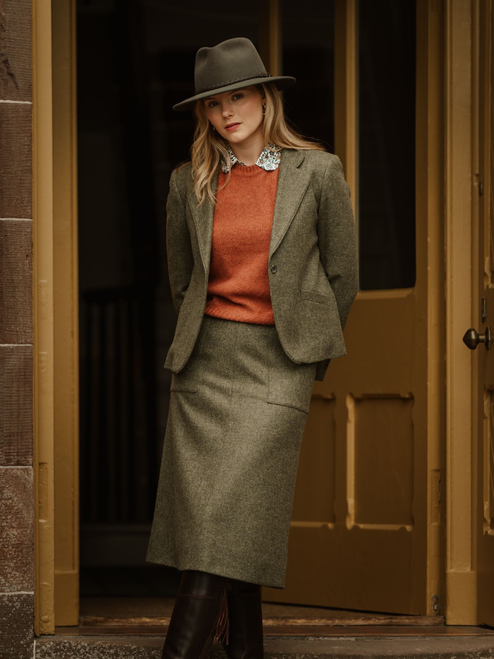 A woman wears a green hat, matching blazer, orange sweater, statement necklace, and the A-Line Skirt by Campbell's of Beauly. She stands before a yellow door, looking at the camera with a neutral expression.