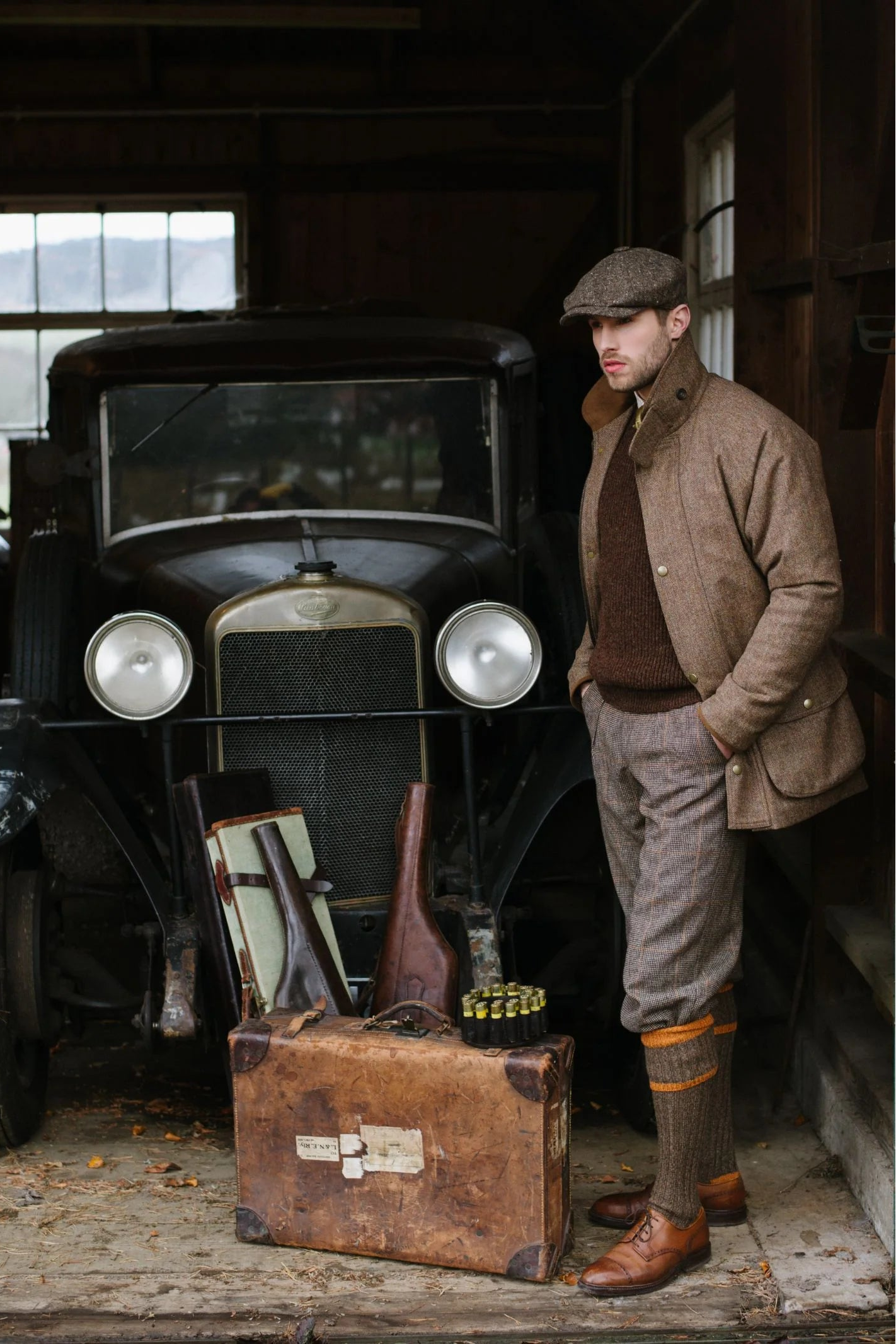 A man in Campbell's of Beauly Plus Fours and a flat cap stands in a garage by a classic car, with a suitcase, two rifles, and an ammo box on the ground before him.