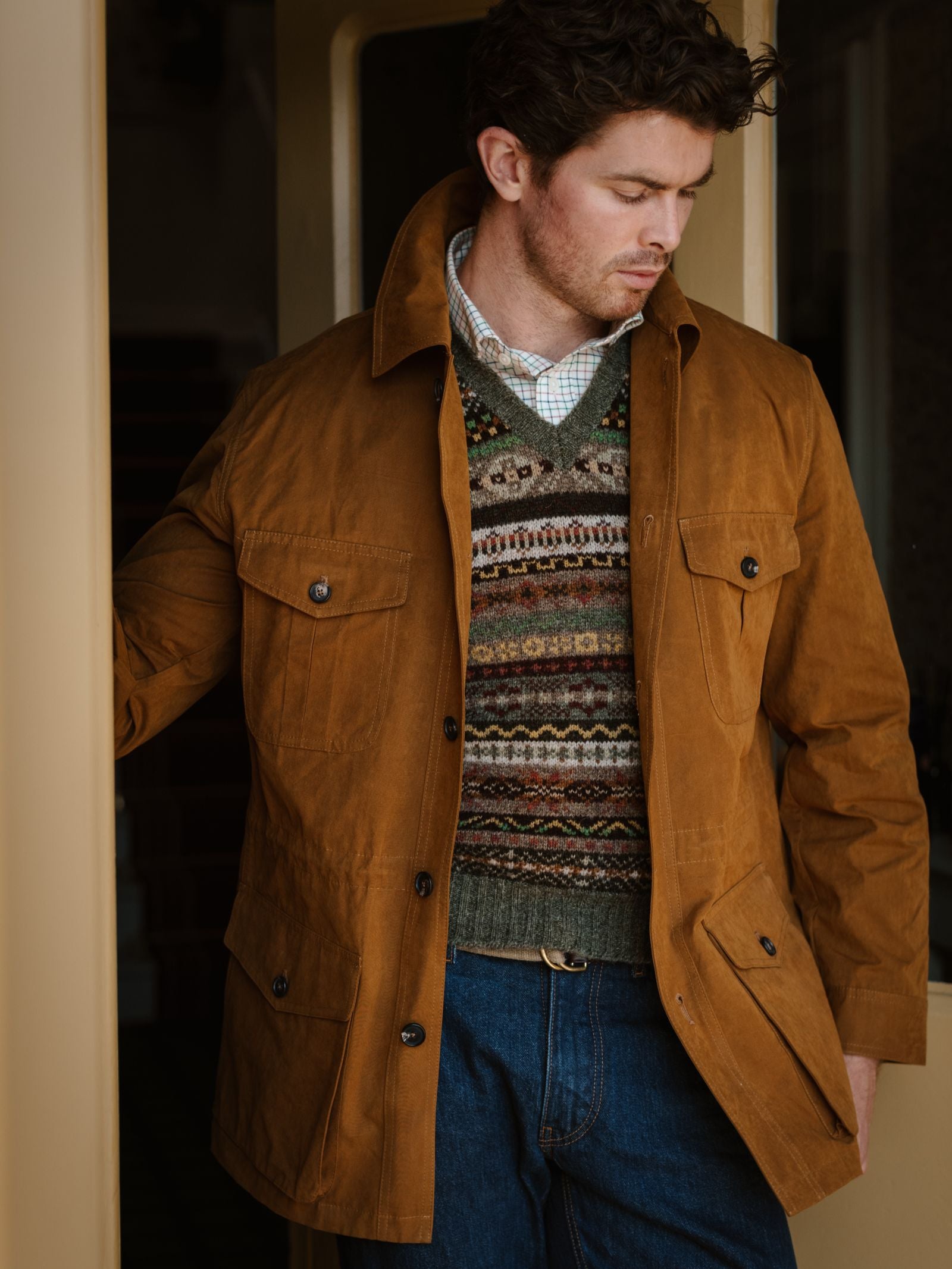 A man in the Aigas Field Jacket by Campbell's of Beauly stands indoors, wearing it over a patterned sweater and plaid shirt with blue jeans, looking down while resting one hand on a door frame. He has short, wavy hair.