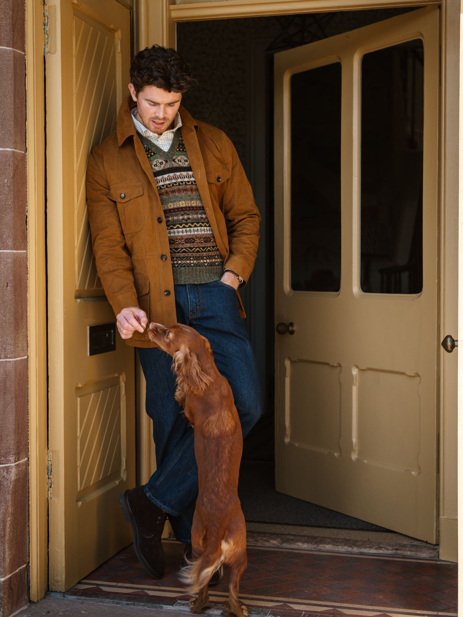 A man in a brown Aigas Field Jacket by Campbell's of Beauly, a patterned sweater, and jeans leans against an open doorway as a brown dog stands on its hind legs reaching up to him.