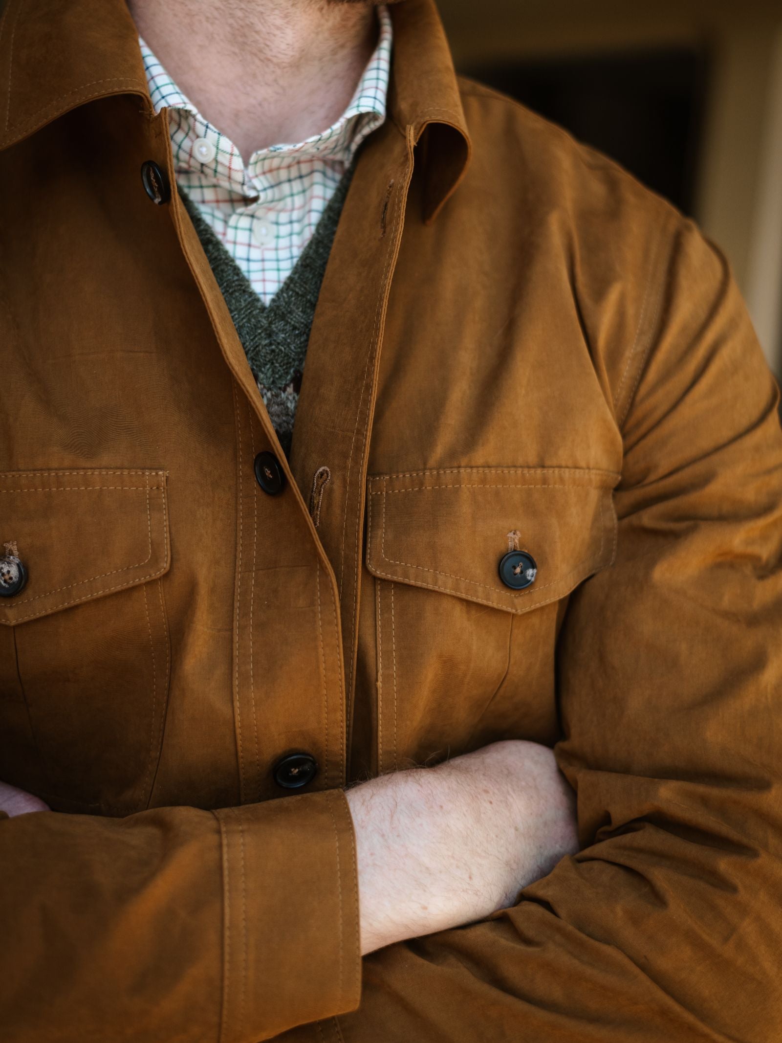 A person with light skin crosses their arms, wearing the Campbell's of Beauly Aigas Field Jacket over a green sweater and white plaid collared shirt. The image is cropped to show only the torso and part of the neck.