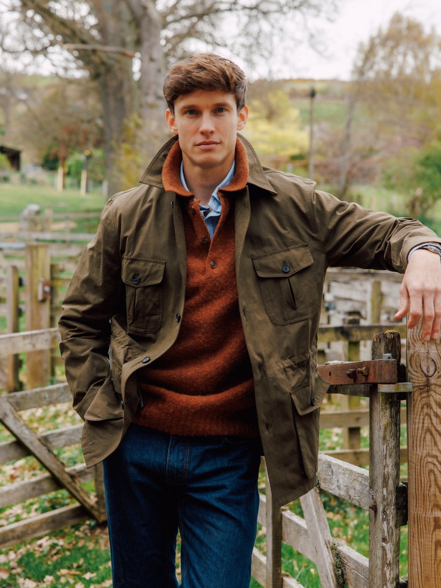 A young man wears the Campbell's of Beauly Aigas Field Jacket with blue jeans as he stands outdoors by a wooden fence, surrounded by green grass and trees on a cloudy day.