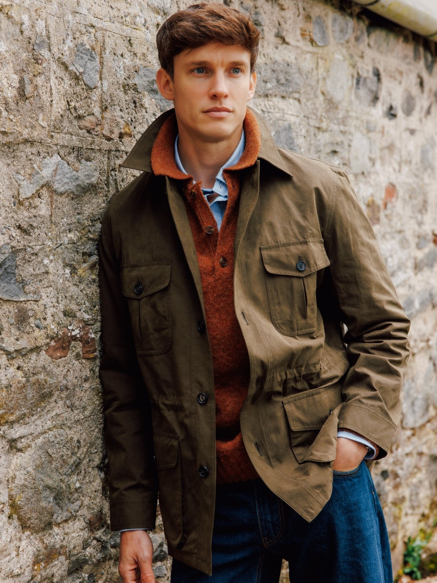A young man with short brown hair stands outdoors by a stone wall, wearing the Aigas Field Jacket from Campbell's of Beauly over a rust sweater and blue shirt with jeans, gazing thoughtfully into the distance.