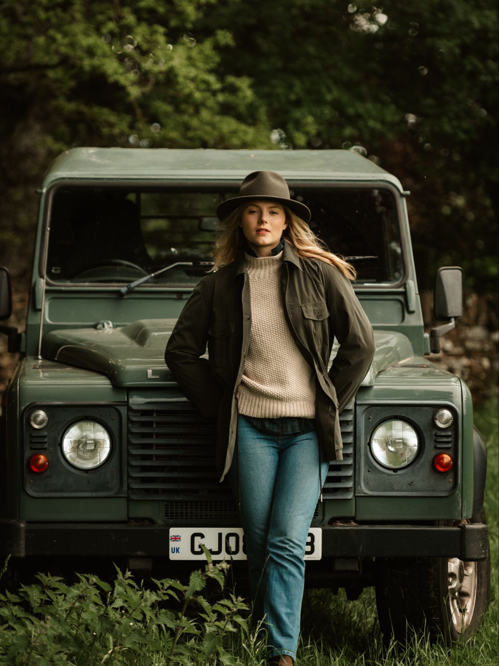 A woman wearing an Australian-made Unisex Akubra Traveller Hat from Akubra, plus a jacket, sweater, and jeans, stands in front of a green vintage Land Rover surrounded by lush greenery.