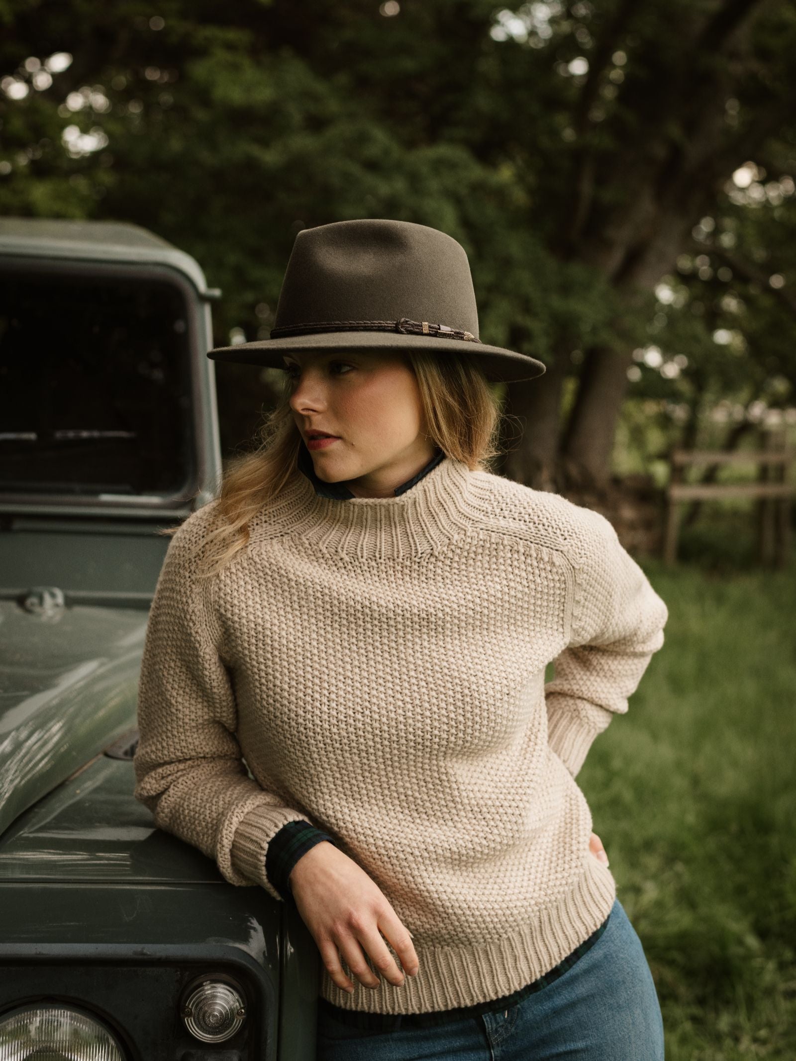 A woman in a beige knit sweater wears an Akubra Unisex Traveller Hat as she leans against a green vehicle outdoors, surrounded by trees and grass. Looking down to her left, she appears thoughtful.