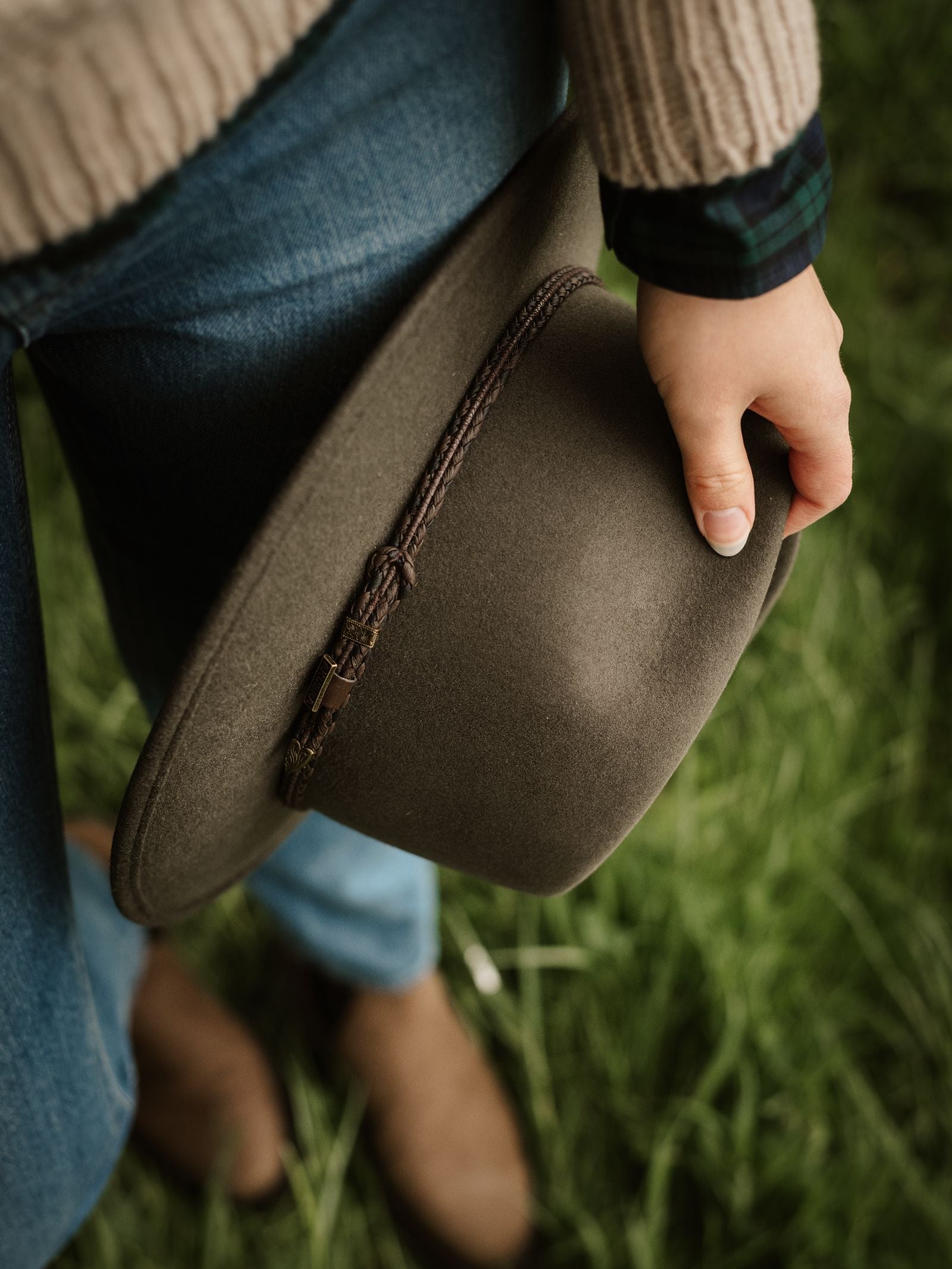 A person in a beige sweater and blue jeans stands on green grass, holding the Akubra Unisex Traveller Hat in Fern by its brown braided band. Only their hand, the hat, and lower body are visible.