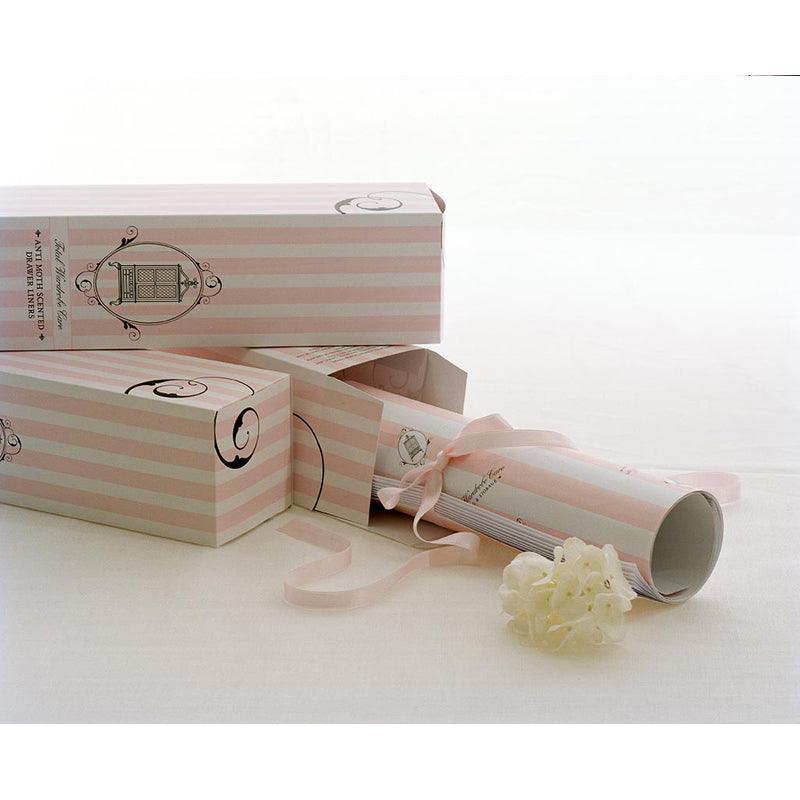 A pastel pink and white striped gift box from Total Wardrobe Care, partially opened to reveal Anti-Moth Drawer Liners tied with a pink ribbon and a decorative white flower, displayed on a plain white surface.