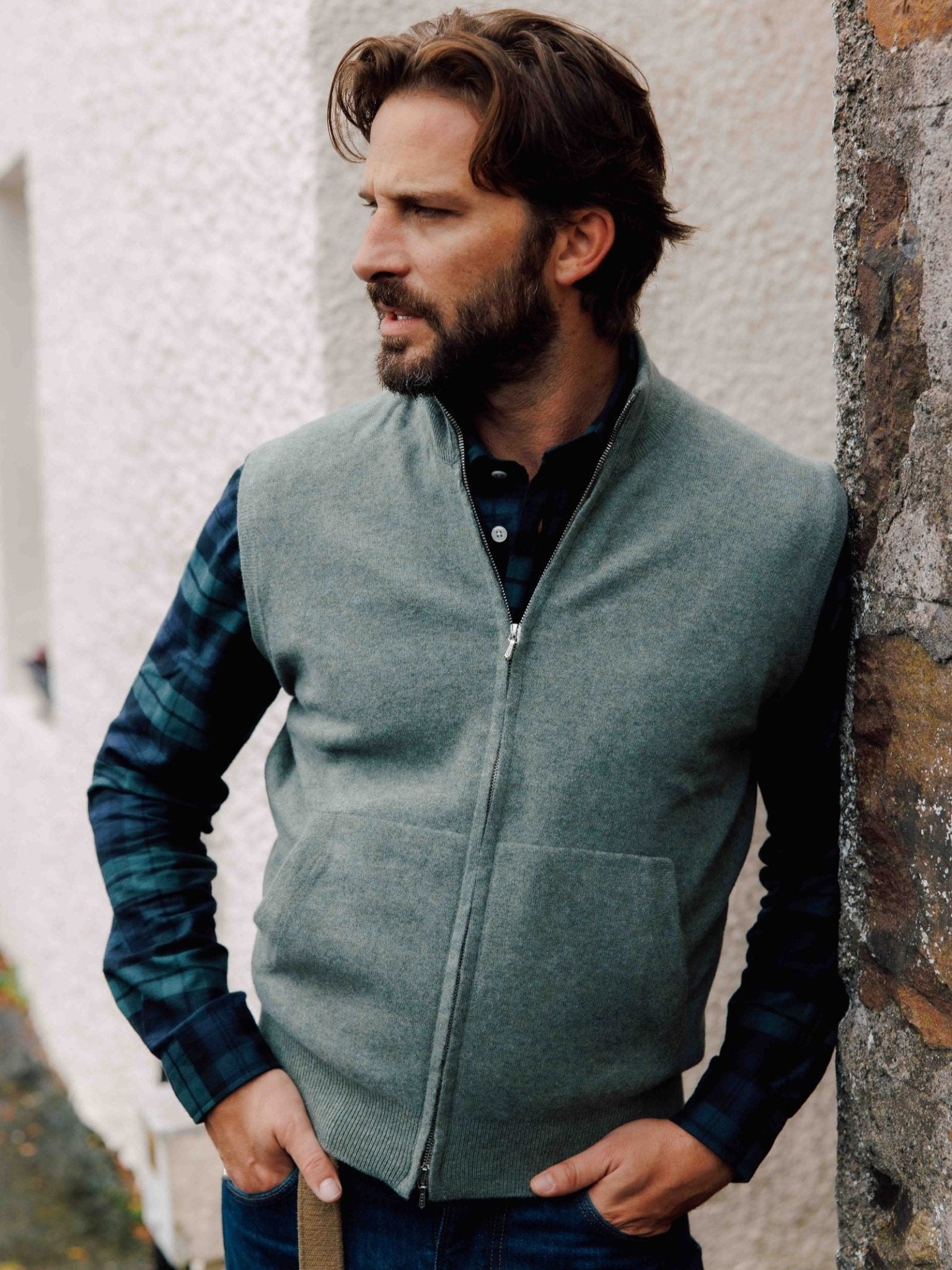 A man with wavy brown hair and a beard stands outdoors in the Scottish Borders, leaning against a stone wall. He wears the Campbell's of Beauly Auld Stock Zip Gilet over a blue plaid shirt, hands in his pockets, looking to his left.