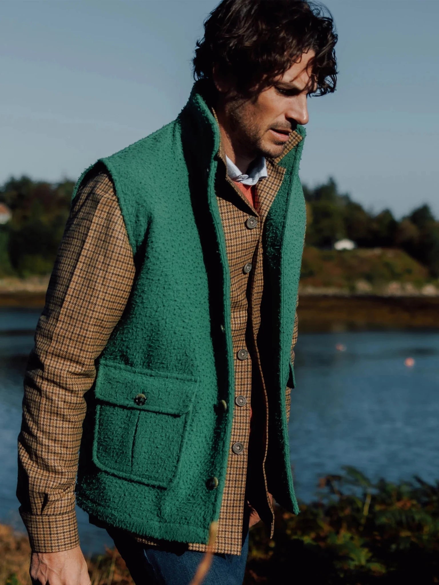 A man with wavy brown hair wears the Campbell's of Beauly Auld Stock Casentino Fleece Gilet in green over a brown plaid jacket, standing outdoors by water with trees and buildings in the background.