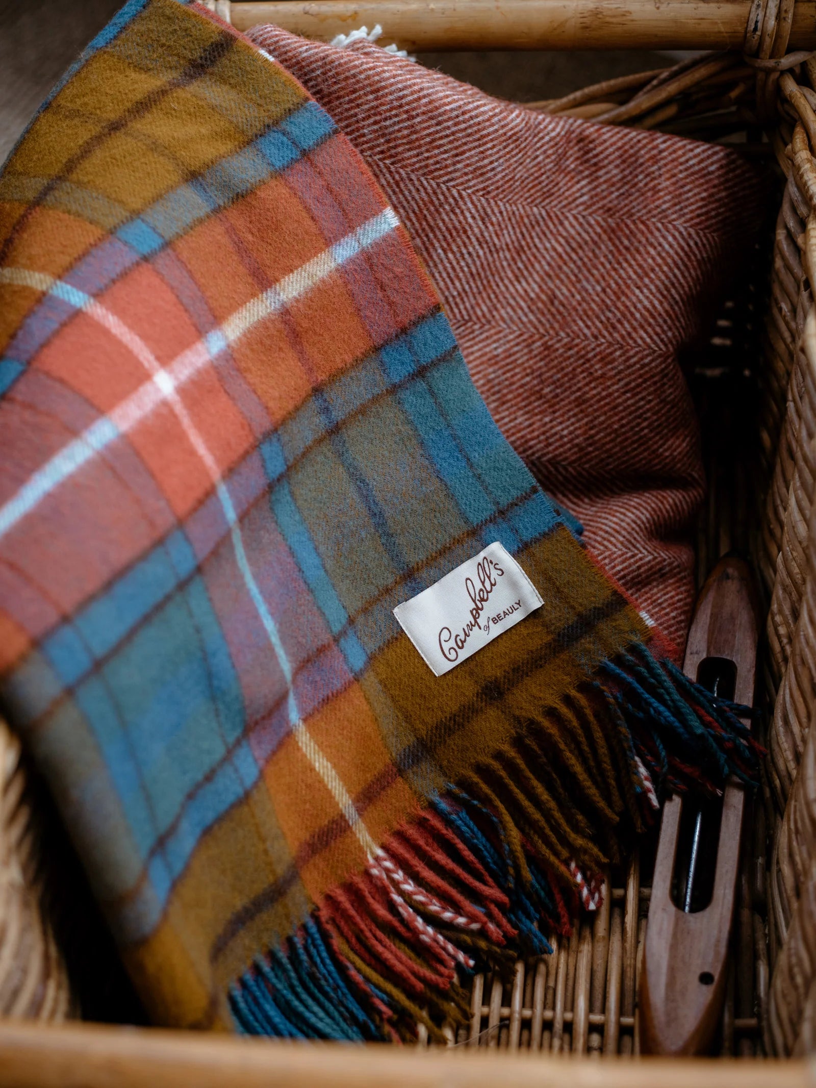 A close-up of a woven basket holds a folded Campbell's of Beauly Lambswool Tartan Blanket, a red herringbone fabric, and a wooden weaving shuttle—highlighting the heritage quality of UK-made throws.