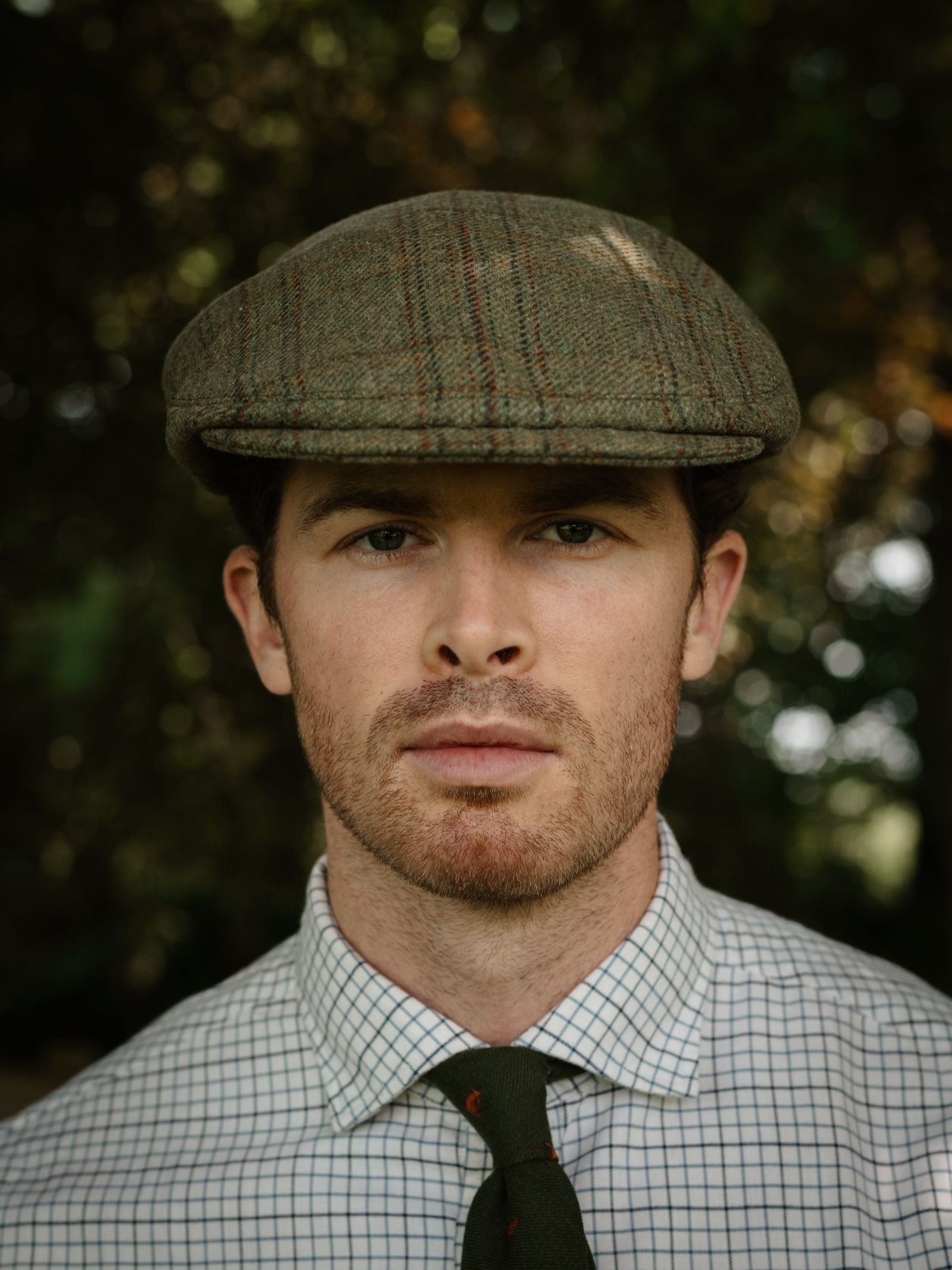 A man with short brown hair and light stubble wears a checkered shirt, green tie, and the Unisex Beauly Cap by Campbell's of Beauly, standing outdoors with blurred greenery in the background.