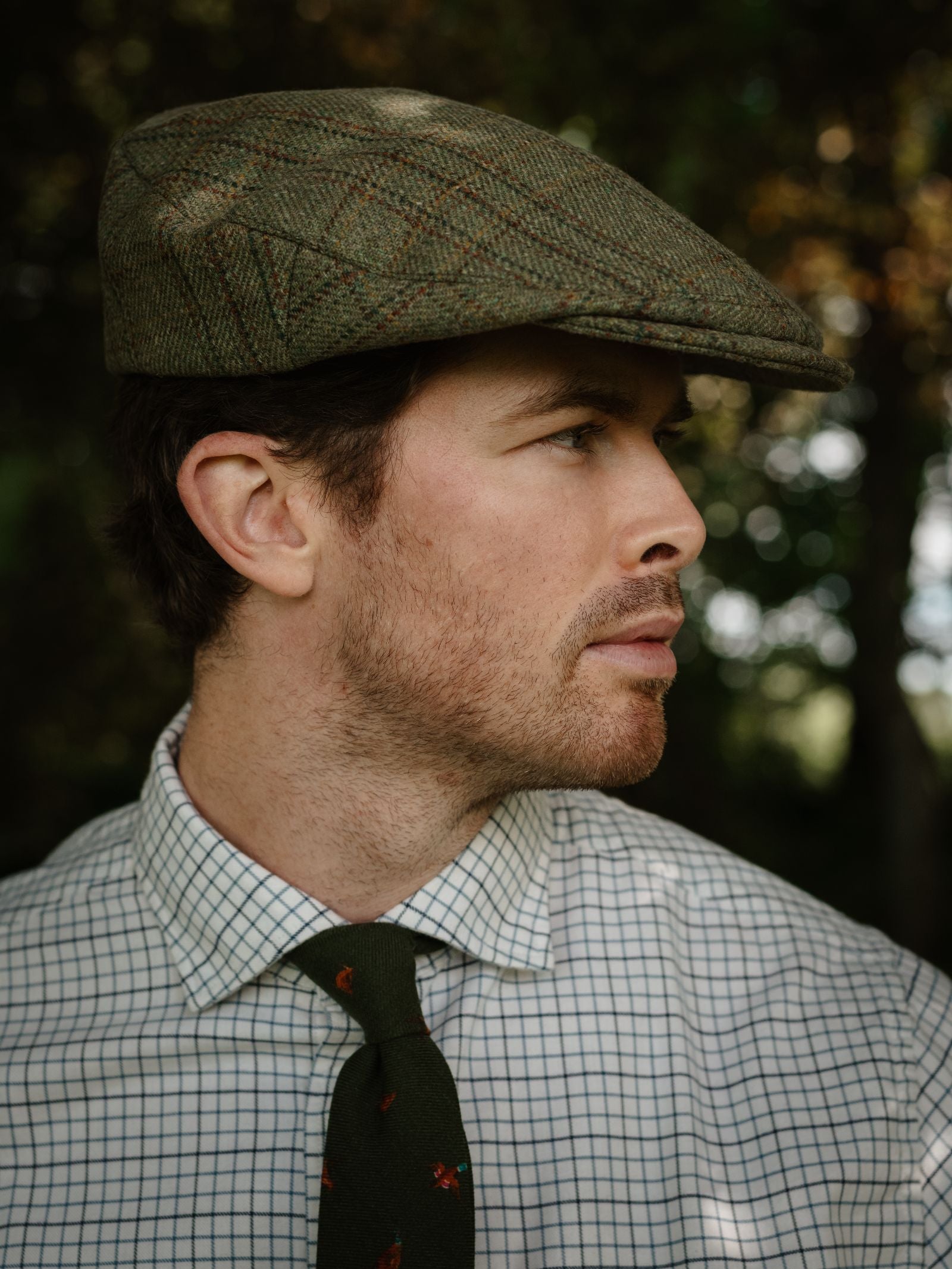 A man with light facial hair wears the Unisex Beauly Cap by Campbell's of Beauly, a white checkered dress shirt, and a dark green patterned tie, standing outdoors with blurred trees in the background.